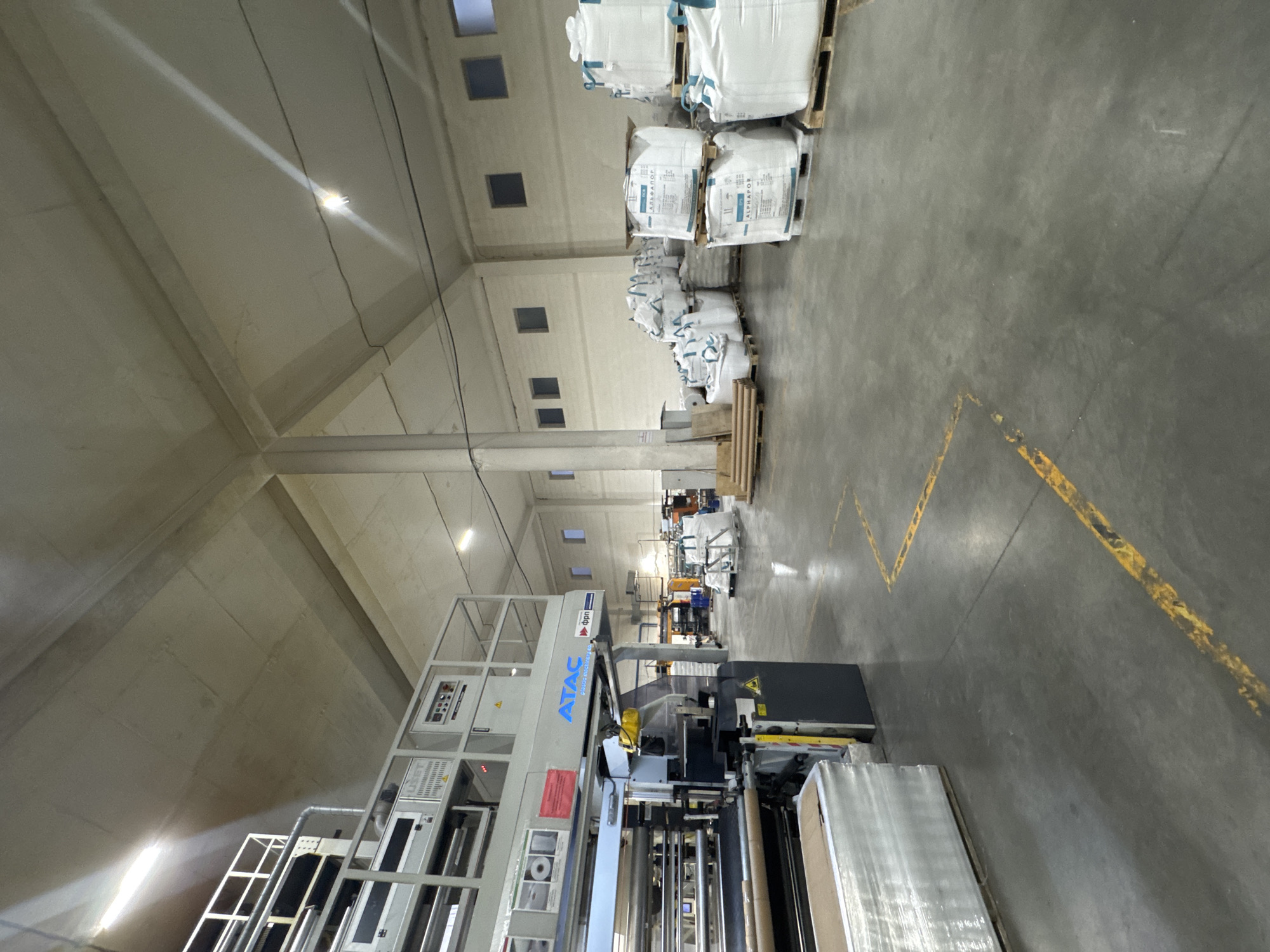 The image shows the interior of a warehouse or factory setting, likely involved in the production or storage of plastic materials. In the foreground, a large piece of machinery, branded "ATAC plastic machinery", is prominent. It appears to be a film extrusion or conversion machine, with various rollers and components visible. To the left of the machine, a pallet of what appears to be stacked boxes, wrapped in clear plastic, is present.
Further into the warehouse, numerous large white bulk bags, also known as FIBCs (Flexible Intermediate Bulk Containers), are stacked on wooden pallets. Some of these bags have labels, with text that is difficult to read clearly but suggests product information or branding. In the background, additional machinery and storage racks are visible, along with several square windows on the far wall. The overall environment is industrial, with a concrete floor marked with yellow lines, and a high ceiling supported by concrete pillars. The lighting appears to be artificial, with bright overhead lights casting long shadows. There are no people visible in the image. The location is identified as Perm, Russia.