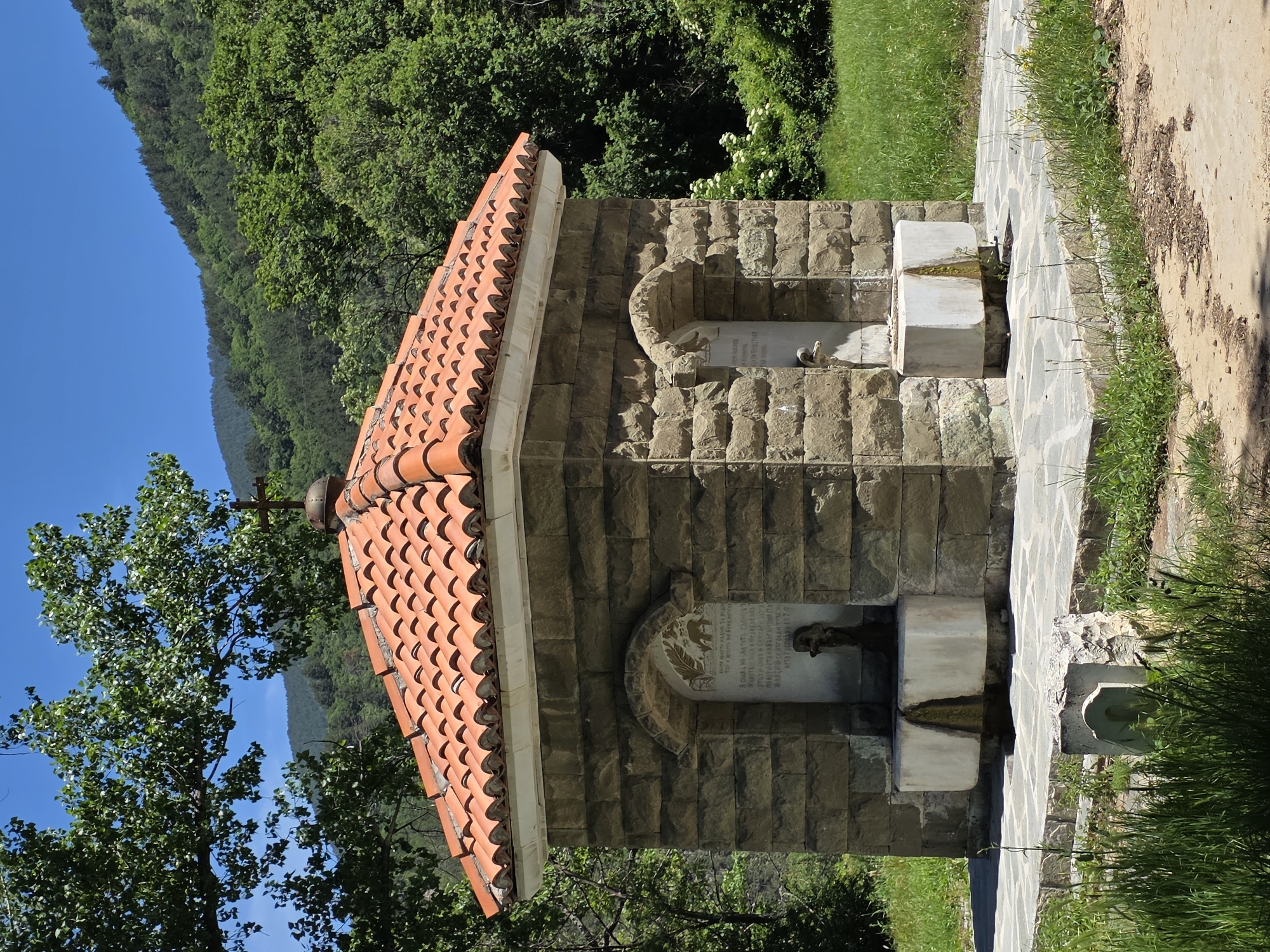 The image captures a stone fountain structure with an orange tiled roof in Bachkovo, Bulgaria. The fountain has two levels, each with a spout for water. The stonework is blocky and weathered, with decorative arches and inscriptions. The background features lush green trees and a blue sky. To the right of the fountain is a stone-paved area with grass and some dirt. A tree is visible on the left side of the fountain. The scene appears to be outdoors in a natural setting near the city of Bachkovo.