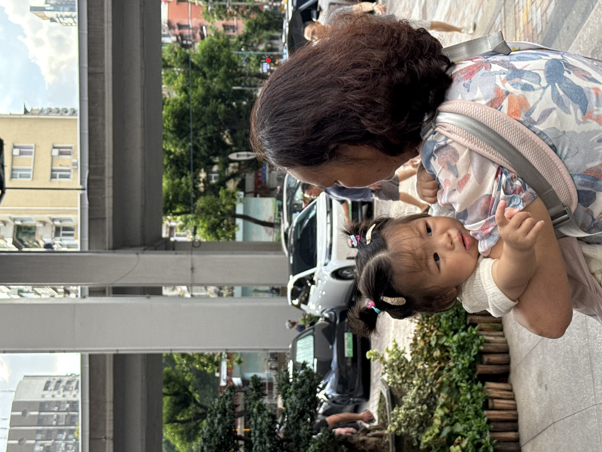 In Haizhu District, China, a woman carries a young child. The woman, with dark, curly hair, wears a floral-patterned shirt and a gray carrier. The child, also with dark hair styled with clips, faces away from the viewer, looking towards the side. They are positioned in the foreground, with a low, wooden-fenced planter box filled with green foliage to their left. In the background, several cars are parked, including a black Tesla and a white car. A pedestrian walkway is visible beneath elevated infrastructure, possibly a bridge or overpass, supported by large concrete pillars. Buildings and trees form the backdrop of this urban scene.