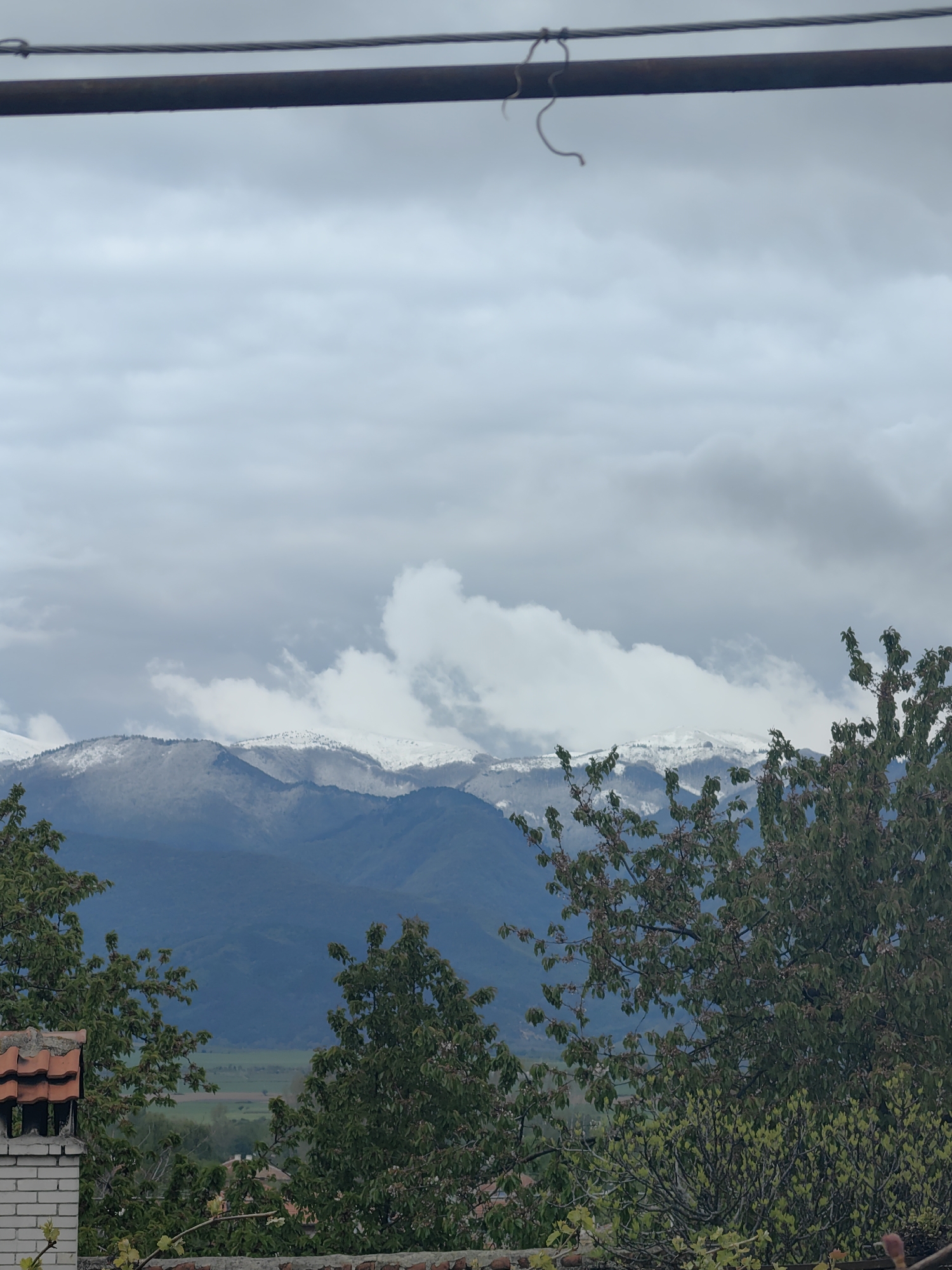 This vertical landscape photograph captures a view of a mountainous region under an overcast sky, likely in Slatina, Bulgaria.
In the background, a range of large mountains dominates the horizon. Their peaks are dusted with snow, contrasting with the darker, forested lower slopes. Patches of white clouds cling to the mountain ridges, partially obscuring them and blending with the gray, cloudy sky above.
The midground reveals a verdant valley, appearing as a mosaic of green fields and potentially distant settlements, stretching between the foreground and the mountain range.
The foreground is framed by lush green foliage. A prominent tree with dense leaves occupies the right side of the image, while another smaller tree is visible on the left. On the lower left, a portion of a brick chimney with orange-red roof tiles is visible, suggesting a traditional building structure. Below the chimney, a weathered brick wall or fence marks the immediate foreground.
Across the very top of the frame, two parallel dark lines, likely utility wires or cables, traverse the sky horizontally. A small, light-colored piece of string or wire hangs loosely from the thinner upper cable.
The sky is heavily clouded with various shades of gray and white, indicating an overcast daytime. The diffused light suggests no direct sunlight. The presence of snow on the distant peaks while the foreground trees are in full leaf points to either early spring or late autumn in a mountainous climate. No human activity or visible text is present.