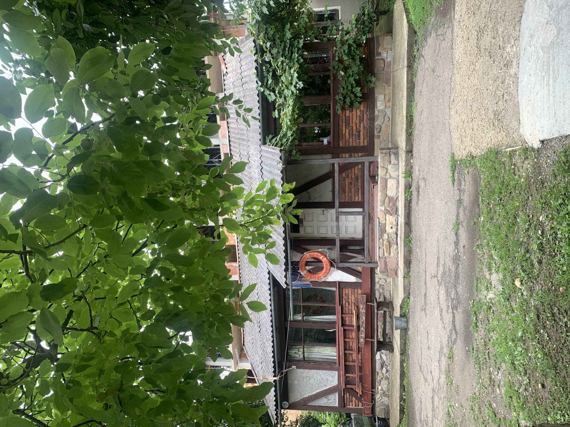 The image depicts a small, single-story building with a prominent pitched roof covered in corrugated asphalt shingles. The front facade features a porch with wooden supports and railings. The lower portion of the building is clad in brick and stone, while the upper section is partially covered in stucco with decorative timber framing. A red life preserver ring is hung on the porch wall. A wooden bench sits in front of the building, next to a metal bucket. The area surrounding the building is paved with asphalt and patches of grass, with a large tree canopy overhead. In the background, a multi-story building under construction is visible. The scene appears to be a residential or recreational property, possibly in a rural or semi-rural setting within the city of Kolomyia, Ukraine.