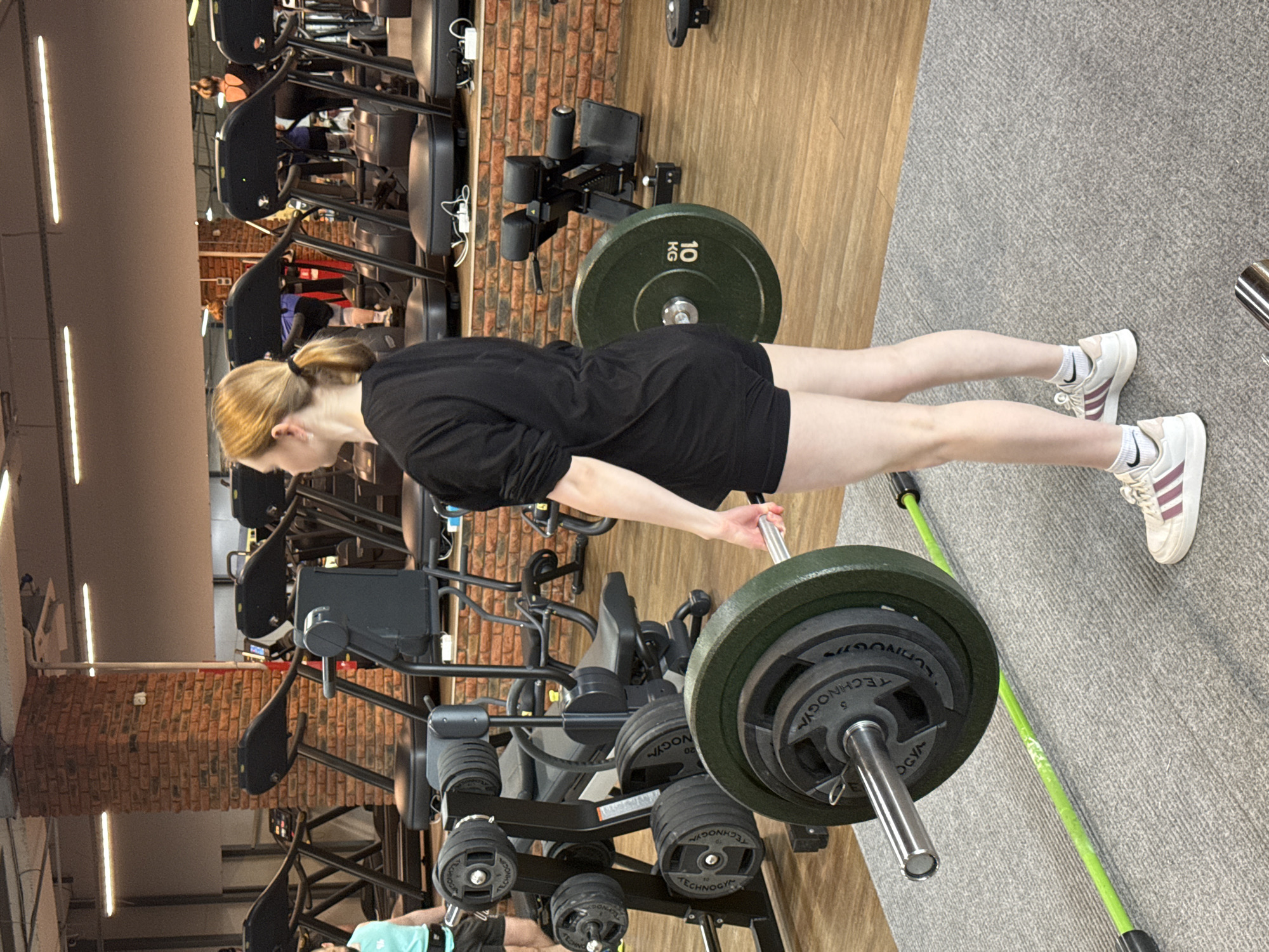 A woman is performing a barbell hip thrust exercise in a gym. She is wearing a black t-shirt and black shorts, white socks and white Adidas sneakers. She is positioned horizontally in the frame.
The barbell has multiple weight plates. One plate is visible and is labeled "10 kg." A green balance stick is lying on the floor beside the barbell. Gym equipment, including treadmills, a weight rack with plates, and other exercise machines, are visible in the background.
The gym has a brick wall and a gray carpet floor. There is no indication that the gym is located in Chaikovsky, Russia, as there are no visible landmarks or signs.