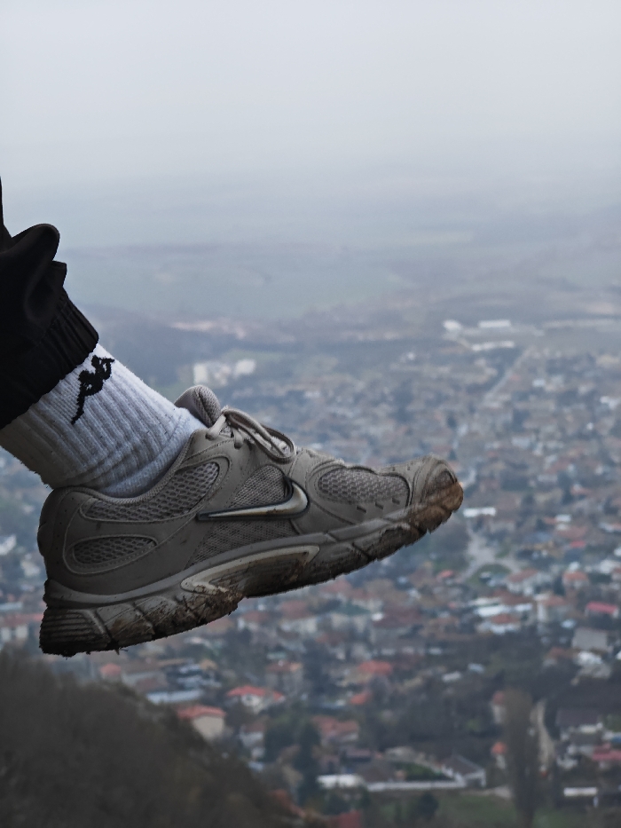 The image presents a close-up, oblique view of a human left foot suspended in mid-air, positioned over a distant urban landscape identified as Shumen, Bulgaria. The foot is clad in a black lower leg garment, a white ribbed athletic sock with a black stylized running figure logo on its lateral ankle, and a light grey/beige Nike brand athletic shoe. The shoe exhibits dried dirt and mud on its sole and forefoot, indicating recent outdoor use. The background features a sprawling cityscape with numerous buildings, many displaying orange and red tiled roofs, consistent with European architecture. Interspersed green spaces and roads are visible within the urban fabric, receding into the hazy distance under an overcast sky. The immediate area below the foot is dark and out of focus, suggesting the edge of an elevated natural formation, such as a cliff or ridge, from which the city of Shumen is observed.