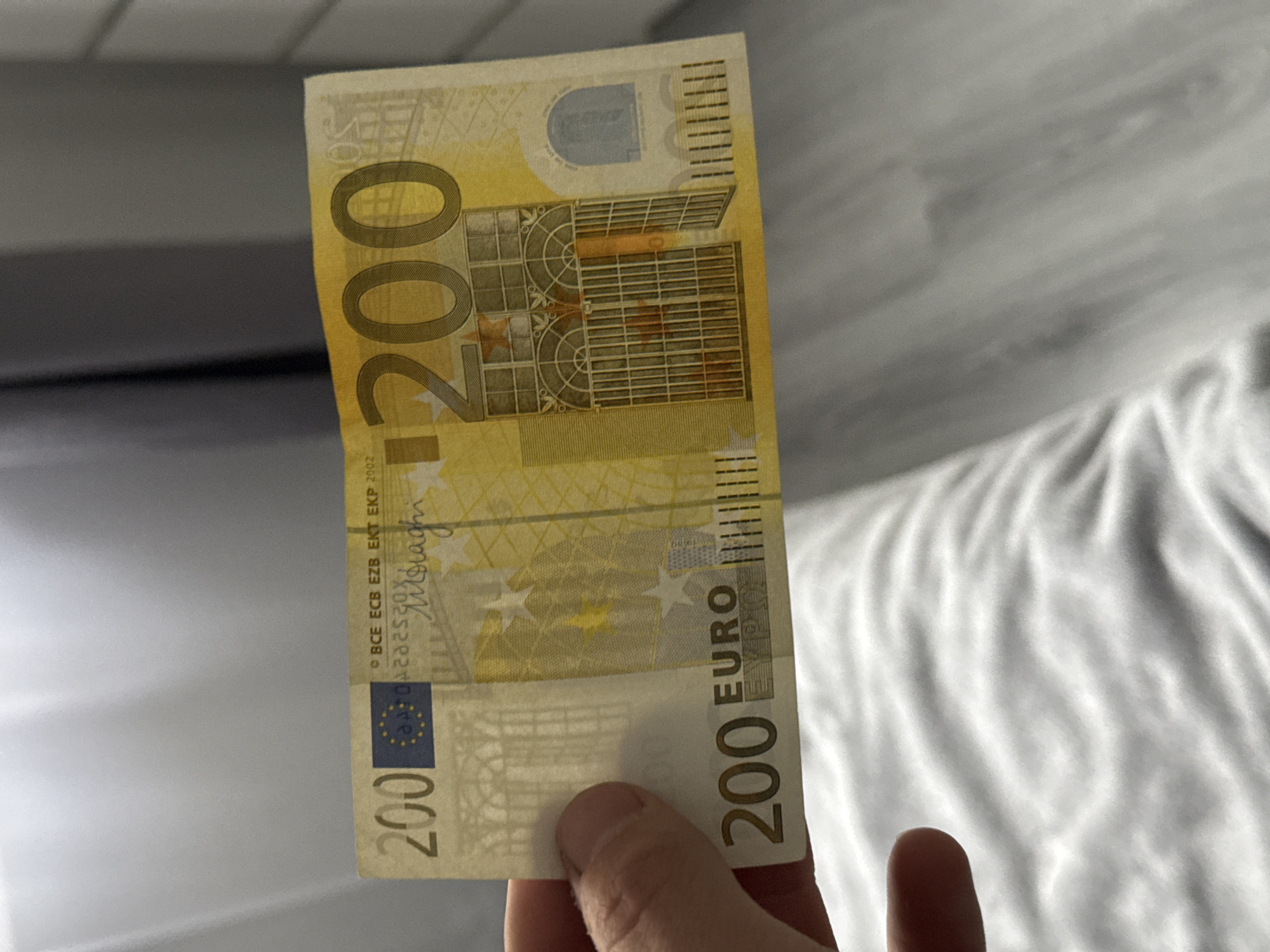 The image displays a close-up shot of a single 200 Euro banknote held vertically by a person's hand. The banknote is predominantly yellow and orange, featuring architectural designs typical of Euro currency, specifically elements inspired by the Baroque and Rococo periods. Large "200" numerals are visible on the left side of the note, with "200 EURO" also printed along the right edge, oriented vertically.
Prominent text details on the banknote include "BCE ECB EZB EKT EKP 2002" and a vertical signature, likely that of a former European Central Bank President. The European Union flag emblem is also visible on the bottom left side of the note. The hand, with visible fingers and thumb, holds the lower portion of the banknote, suggesting it is being presented or examined.
The background is softly blurred. To the left and upper part of the image, a darker, possibly gray or black, smooth surface is visible. To the right and lower part, there is a lighter, white or off-white fabric, appearing to be a rumpled bedsheet or similar soft material, creating a textured contrast with the crisp banknote. The lighting is diffused and even, indicating an indoor setting. The image captures a simple moment of handling currency. The location context provided is Stara Zagora, Bulgaria.