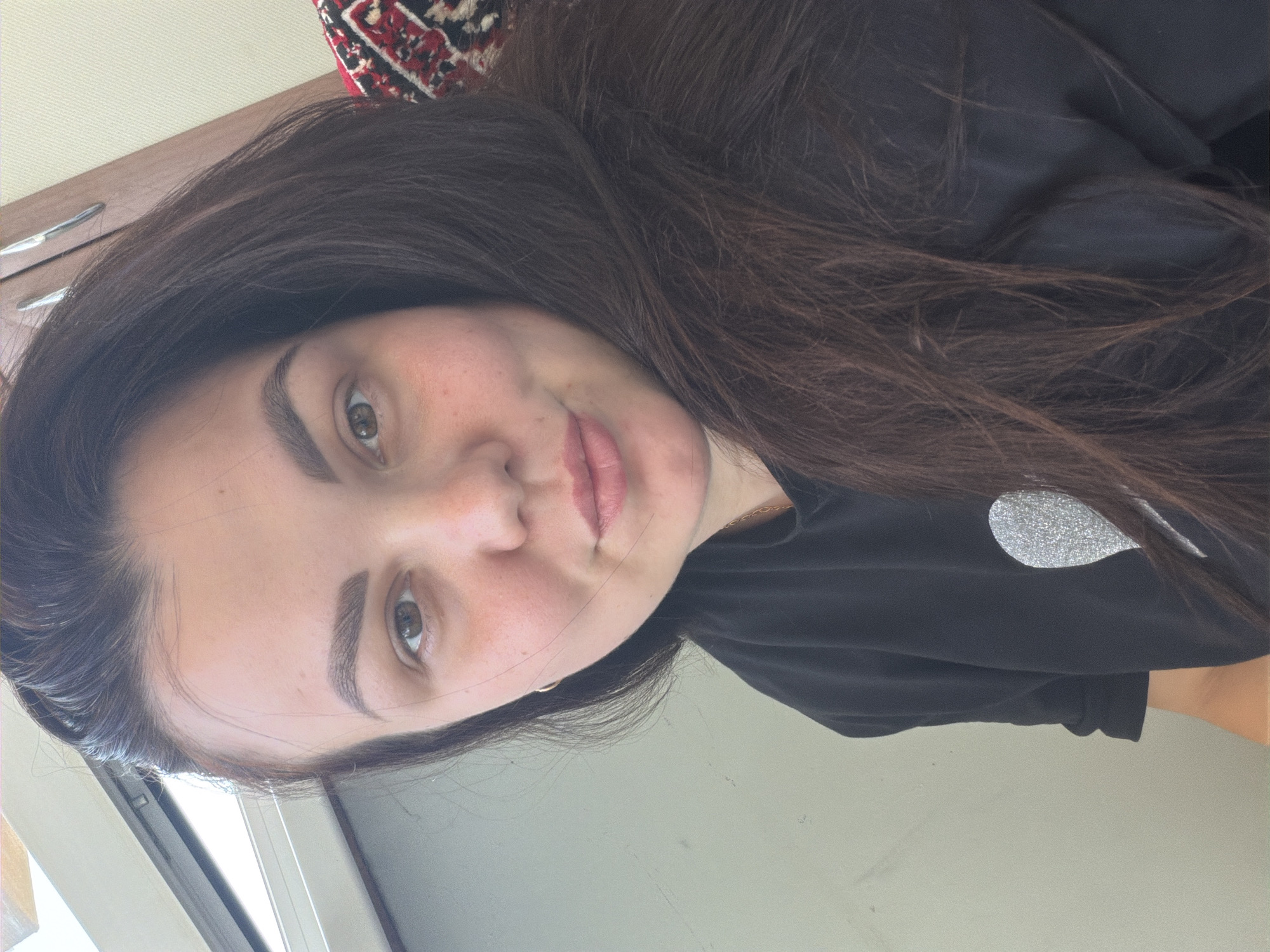A close-up, self-taken photograph shows a young woman with long, dark brown hair. She has light skin with some freckles and a hint of blush on her cheeks. Her eyebrows are dark and well-defined, and her eyes are a warm brown. Her lips are slightly parted, and she has a subtle smile. She is wearing a black t-shirt with a shimmering, silver heart-shaped design on the left side of her chest. The background is out of focus, but appears to be indoors with natural light coming from a window on the left. A patterned textile, possibly a blanket or cushion, is visible on the right side of the frame. The image does not provide specific details about the city of Izhevsk, Russia.