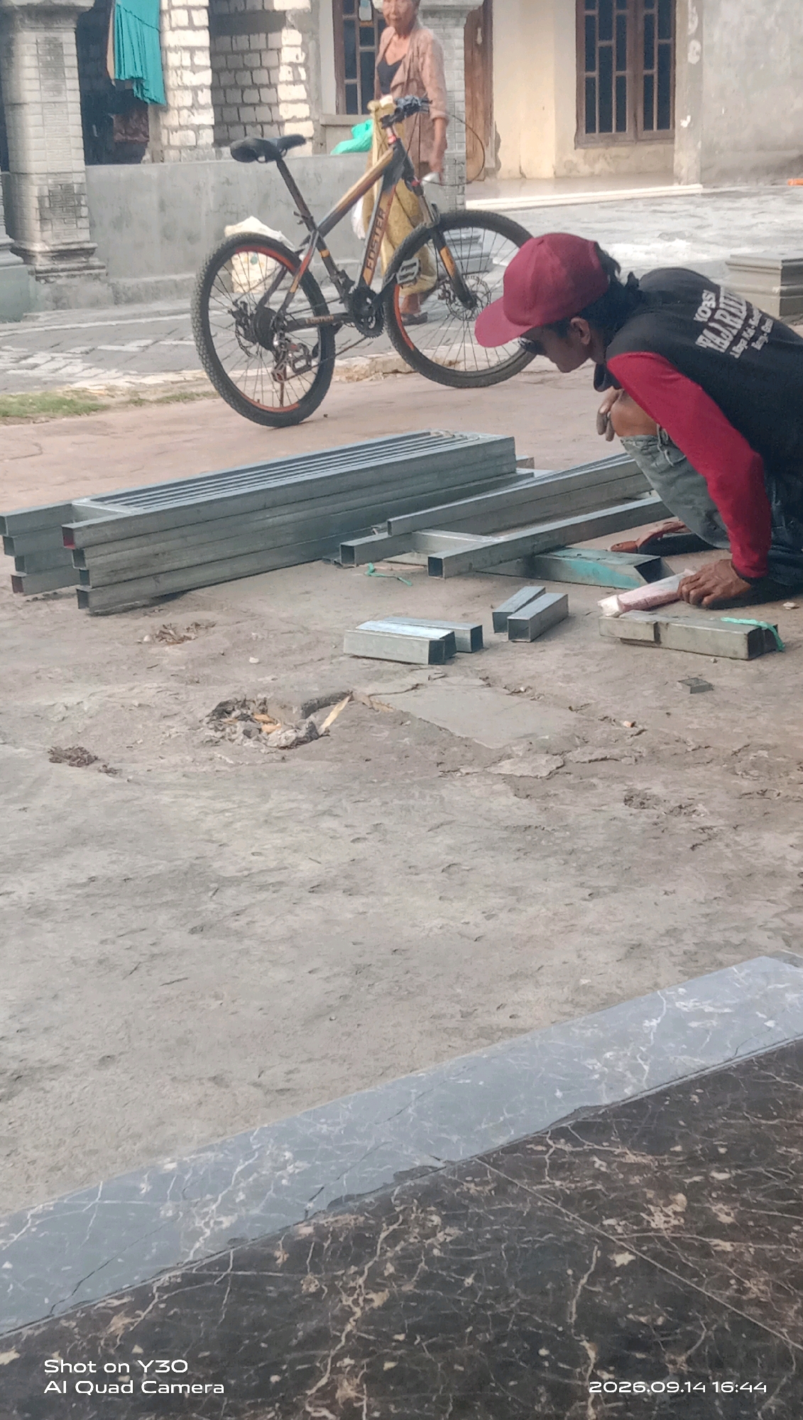 An elderly woman in a light-colored, patterned shirt and pants stands near a bicycle. A young man wearing a red cap and a black shirt with red sleeves is crouching on the ground. He appears to be working with metal pipes, which are neatly stacked and laid out around him. The scene takes place outdoors on a paved surface, likely a street or yard, with buildings visible in the background. The image also shows a section of a marble-patterned surface in the foreground with text overlays indicating it was "Shot on Y30 AI Quad Camera" and "2026.09.14 16:44". The location is identified as Karangankidul, Indonesia.