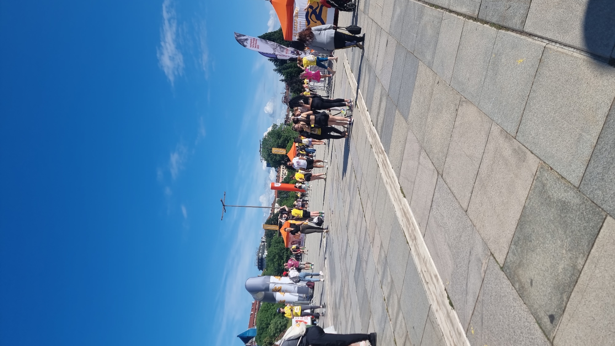 A sunny daytime outdoor event is taking place on a large paved plaza, likely in Sofia, Bulgaria, given the context of the image. The sky is bright blue with a few scattered clouds. In the background, there are buildings and trees. The foreground and midground are occupied by a crowd of people gathered for an event, possibly a race or festival.
Several event-related structures are visible. There are large inflatable arches, tents, and banners, some of which display text and graphics. The people are dressed casually, many in athletic wear or t-shirts. Some individuals appear to be participants, while others might be spectators or event staff. There are groups of people interacting, and some are walking or standing around.
The plaza itself is made of large, square stone pavers. A low, lighter-colored border runs through the paving stones, creating distinct sections. The overall atmosphere suggests a public gathering or a sporting event.