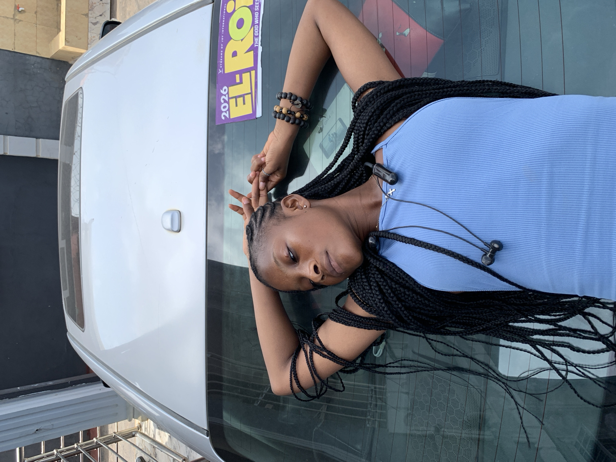 A young woman with braided hair is lying on the back of a white vehicle. She is wearing a blue ribbed tank top, black earphones, and bracelets made of wooden and black beads. Her right hand is raised in a peace sign. The vehicle has a sticker that reads "EL-ROI 2026 STREAMS OF JOY INTERNATIONAL THE GOD WHO SEES." The location is identified as Ikorodu, Nigeria.