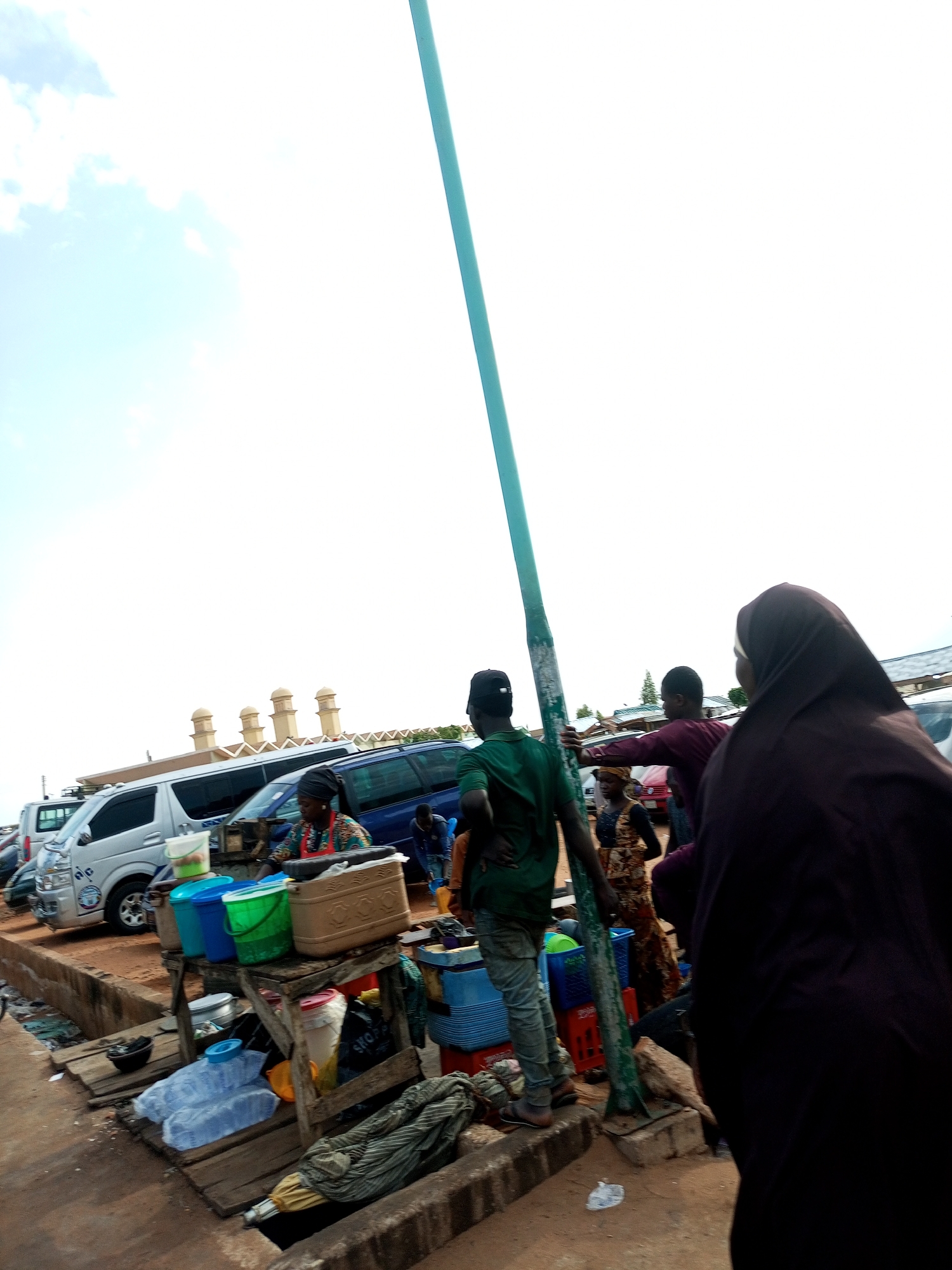 An outdoor market scene unfolds with a bright sky overhead. In the foreground, a tall, light blue pole stands prominently. To the right, a person wearing a dark purple hijab and robe is visible from the back. Further to the left, a vendor with a headscarf and a patterned dress stands behind a wooden table laden with various items, including buckets, containers, and what appear to be food products. Several vehicles, including vans and cars, are parked in the background, suggesting a busy commercial area. A woman is seen tending to her stall, while other individuals mill about the market. The ground is made of dirt and concrete.