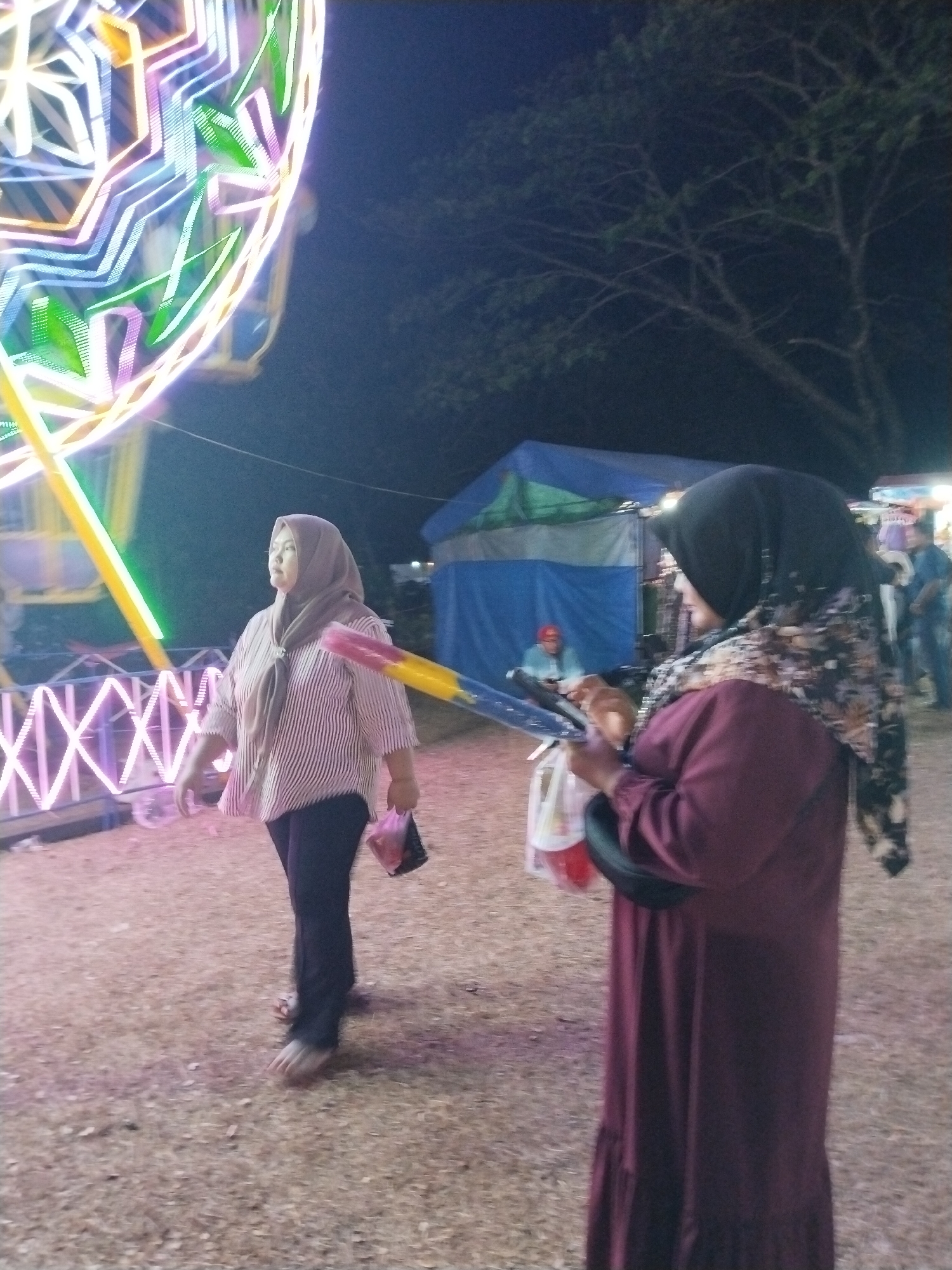 In Tasikmadu, Indonesia, a nighttime scene unfolds at what appears to be a fair or carnival. The background is dominated by a large, brightly lit Ferris wheel, its geometric patterns illuminated by a spectrum of colors, predominantly white, green, and pink. The ground is a rough, earthy surface, with scattered dry leaves and debris.
In the foreground, two women are the primary subjects. The woman on the left, wearing a light brown hijab and a horizontally striped long-sleeved shirt in shades of pink and white, is walking from left to right, her gaze directed upwards and slightly to her right. She holds a pink and black plastic bag in her right hand. Her dark pants and sandals are visible.
The woman on the right, wearing a black hijab with a floral pattern and a long, deep red dress, is facing away from the camera, looking to her left. She is holding a colorful cylindrical object, possibly a toy or decoration, in her hands, along with a plastic bag. A black bag or accessory is slung over her left shoulder.
Behind the women, several stalls or tents are visible, one with a blue and green canvas roof. The atmosphere is dimly lit, characteristic of an outdoor evening event. The overall impression is that of a lively gathering at a local fairground.