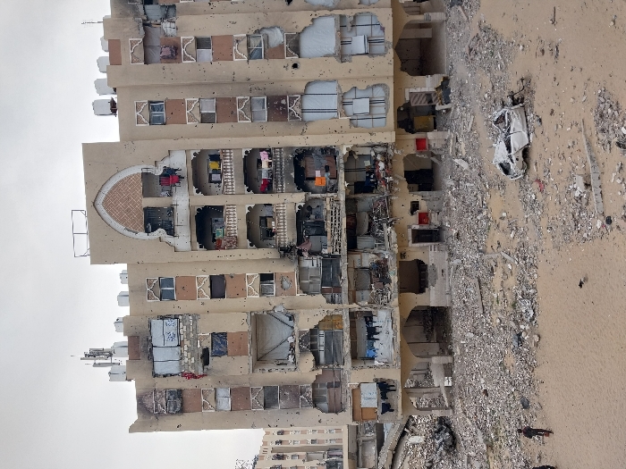 An eight-story residential building in Khan Yunis, Palestinian Territory, exhibits extensive damage under an overcast sky. The building's cream-colored facade is riddled with numerous bullet and shrapnel holes, and multiple sections of walls are compromised or missing, exposing interior rooms. Many windows are shattered or boarded with makeshift coverings, while some balconies retain clotheslines with laundry, including red, yellow, and darker garments. Communication antennas and water tanks are visible on the roof, some appearing damaged.
The ground in front of the building is covered in a dense layer of concrete rubble, rebar, and debris. A heavily damaged, crumpled white sedan is positioned on a patch of sandy ground to the right of the frame. Two individuals, appearing to be children or young adults, walk across the sandy foreground towards the left, away from the main structure. A second, similarly damaged multi-story building is partially visible in the distant background on the left.