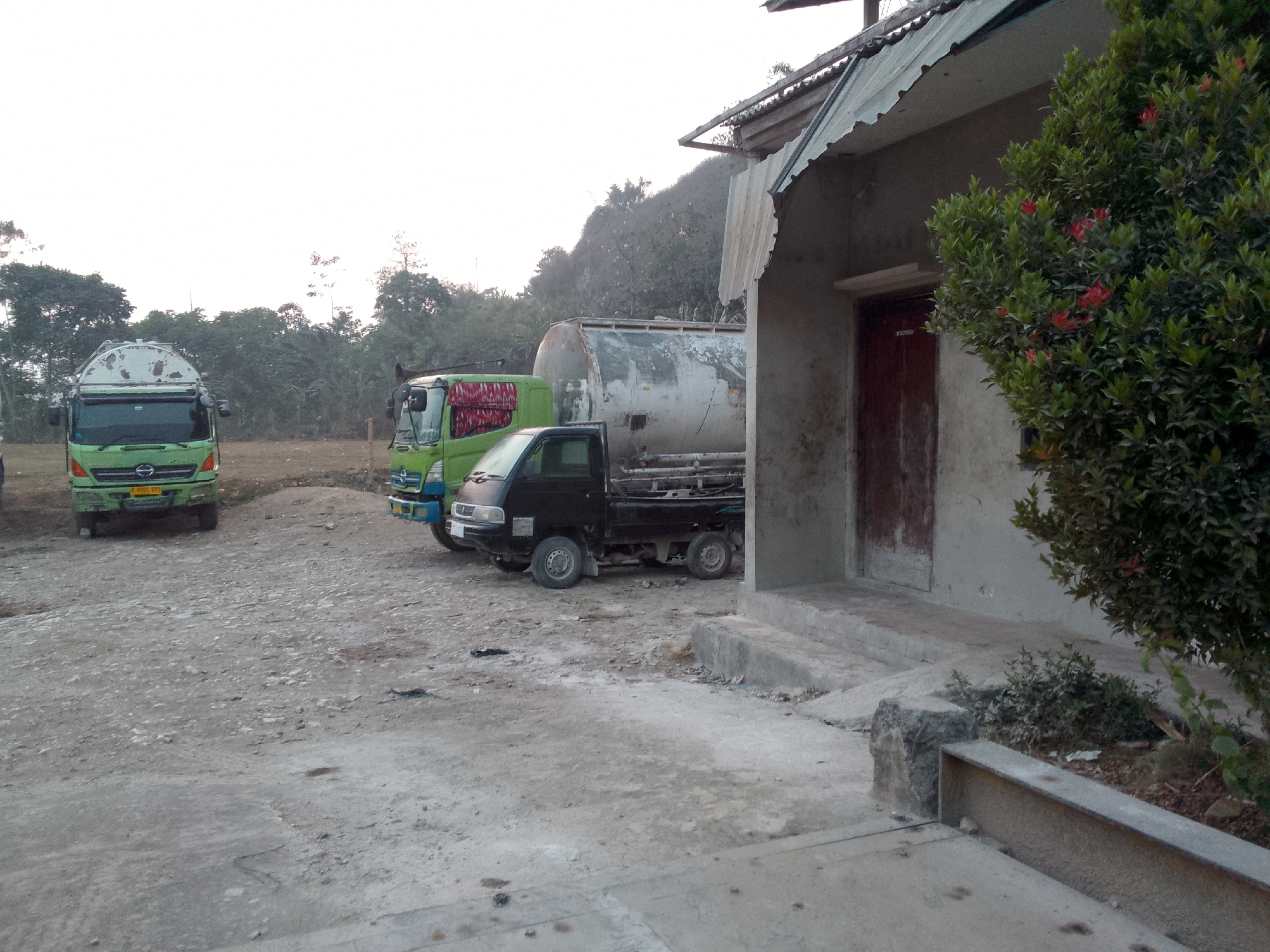 The image depicts a scene in Gunungmasigit, Indonesia, featuring a parked collection of vehicles and a building. In the foreground, a dusty, gravelly lot is visible, leading to a building with a corrugated metal roof and a wooden door. Adjacent to the building, a dense green shrub with red flowers is in bloom.
To the left of the building, several trucks are parked. The closest truck to the building is a small black pickup truck. Behind it, a larger green truck with a white tanker body is parked. Further to the left, another green truck, this one with a lighter colored cab, is parked. The environment appears to be an outdoor staging area or a parking lot, possibly near a factory or industrial site, given the presence of the trucks and the utilitarian nature of the building. The sky is overcast, suggesting early morning or late afternoon lighting conditions. No people are visible in the image.