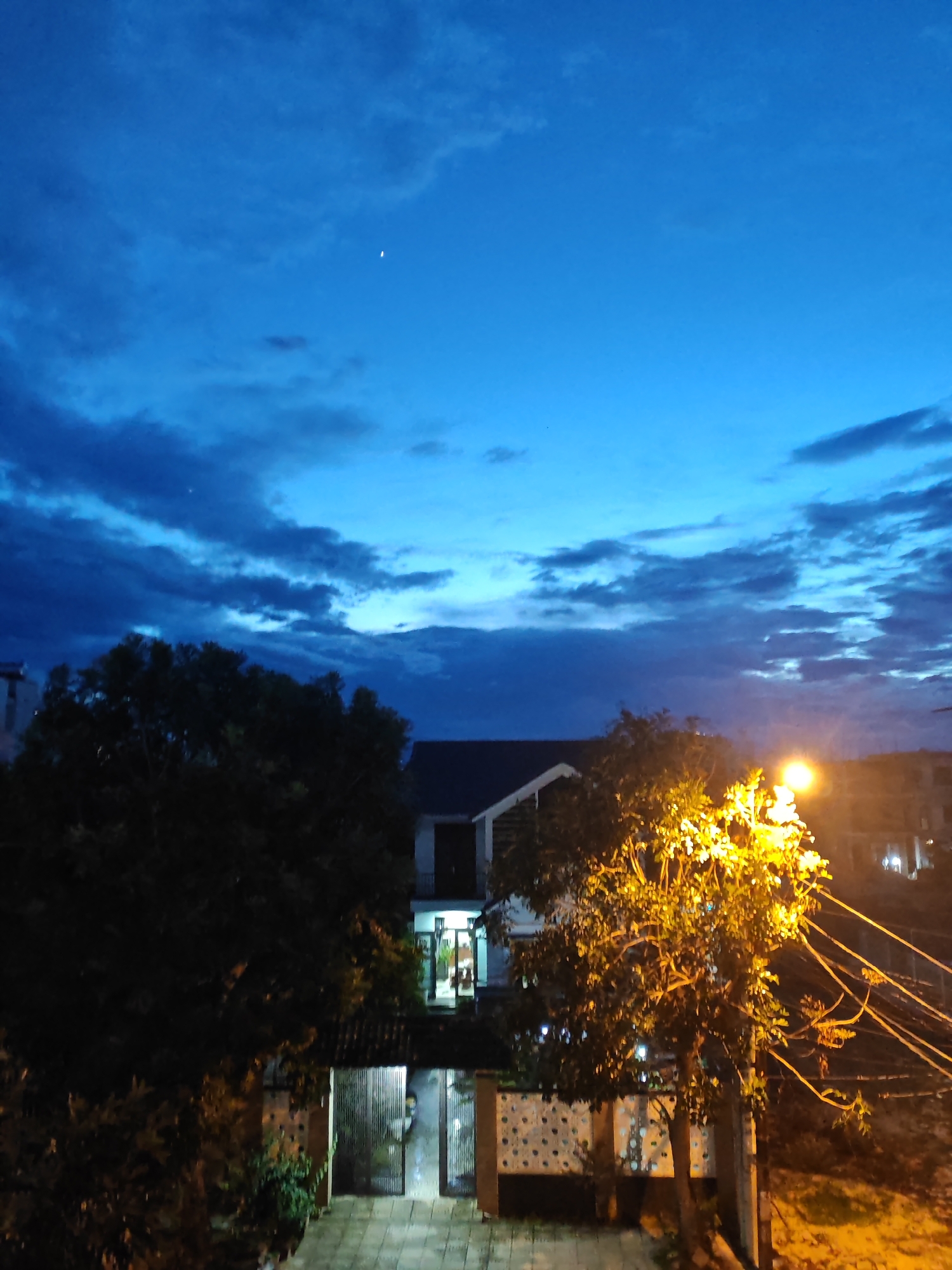 The image shows a twilight scene in Đà Nẵng, Vietnam. A bright streetlamp illuminates a tree and power lines in the foreground, casting a warm glow. Behind it, a residential building with interior lights on is visible, with an open gate and courtyard. Dark trees frame the left side of the image, and in the distance, other buildings are faintly visible. The sky is a deep blue with scattered clouds, and a sliver of the crescent moon is visible near the top. The overall impression is of a peaceful evening in a residential area.
