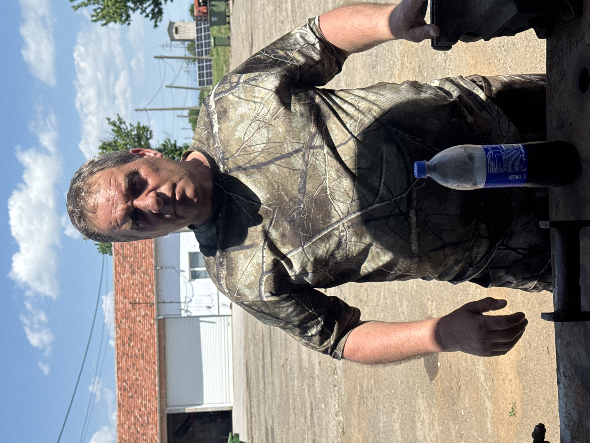 A middle-aged man in a camouflage shirt stands outdoors, looking towards the camera. He appears to be working on or interacting with a piece of machinery. A bottle of dark liquid sits nearby. The setting is Sarnevo, Bulgaria, as indicated by the visible architecture and general environment. Behind the man, a building with a red brick roof and a white garage door is visible. The sky is blue with scattered clouds. In the distance, utility poles, solar panels, and a small structure are discernible. The ground is concrete, suggesting an industrial or agricultural setting.