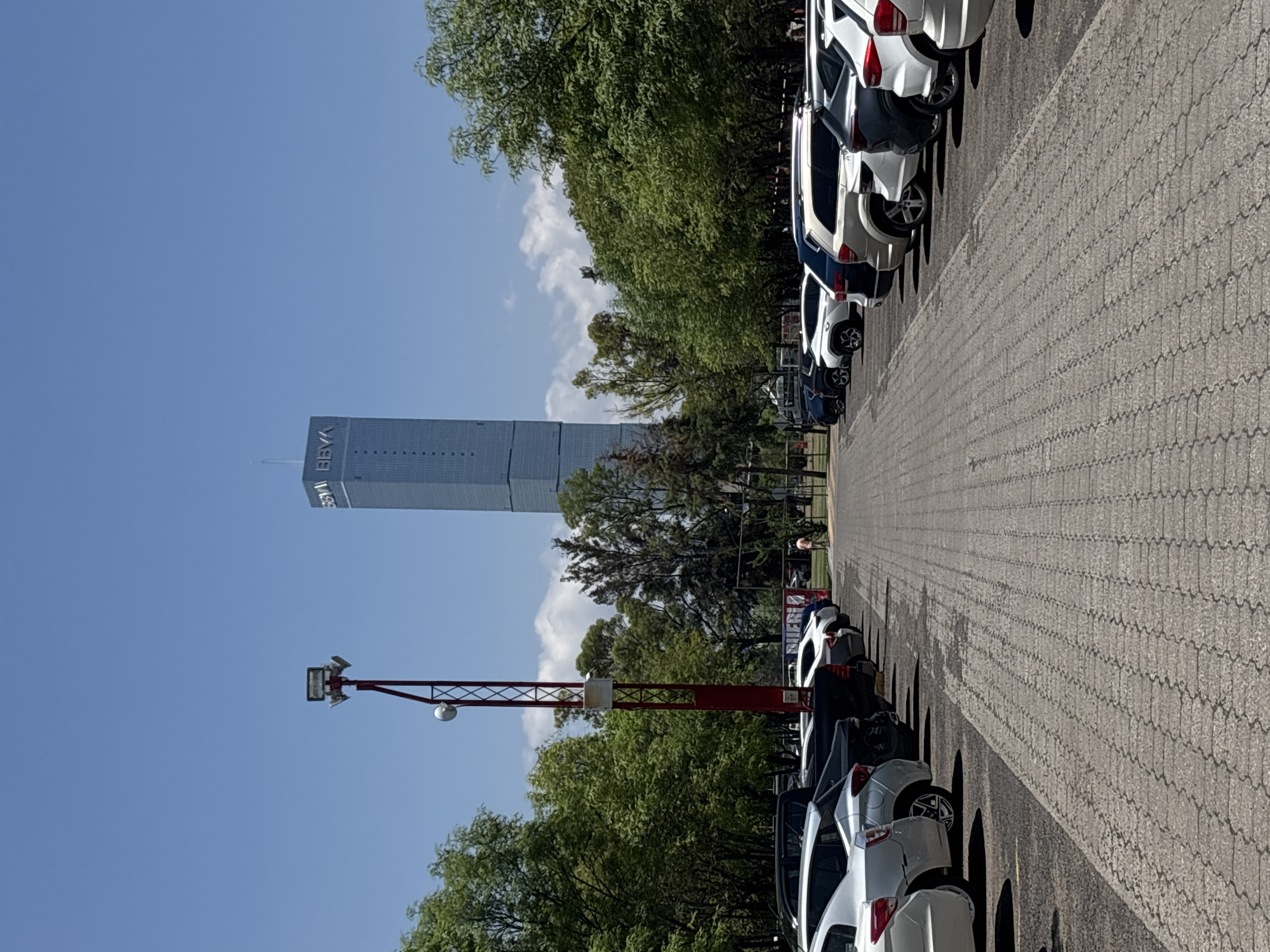 The image depicts a parking lot in San Bernardino Tlaxcalancingo, Mexico, with a row of parked white and dark colored vehicles. The BBVA tower rises in the background. A red metal structure with mounted lights stretches across the foreground. The sky is clear and blue with a few clouds. Green trees are present along the side of the parking lot. The parking surface is paved with interlocking gray bricks.
