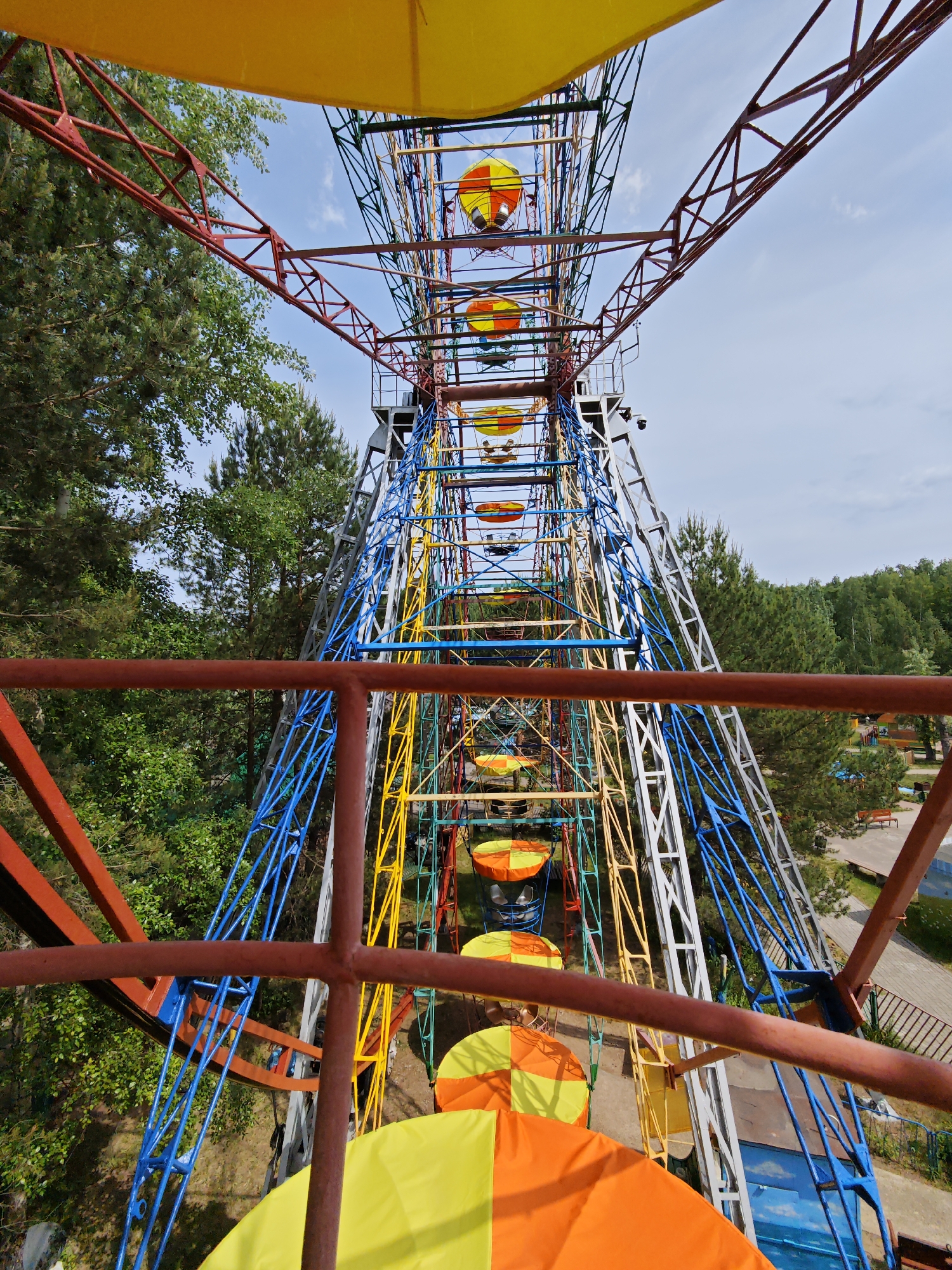 The image shows a Ferris wheel in the city of Chaikovsky, Russia. The view is looking up and through the wheel's structure. The Ferris wheel has a multi-colored frame with red, blue, yellow, and silver support beams. The passenger cars are orange and yellow. The backdrop includes green trees and some blue sky. There is red metal railing in the foreground.