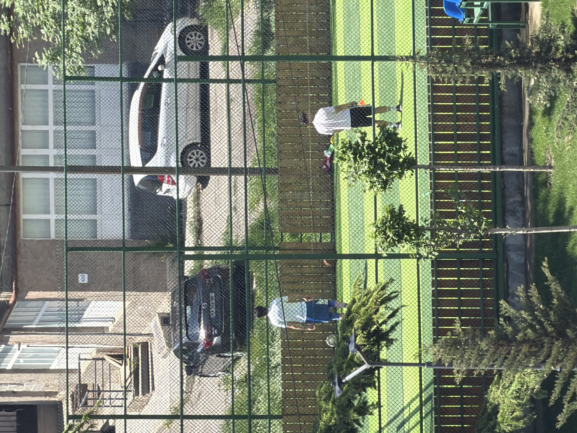 From an elevated perspective behind a green chain-link fence, the scene includes a residential building, a street, a sports court, and people. The location is Tbilisi, Georgia, as evidenced by the license plate on the dark-colored car, which reads "XX-362-XX GE."
The residential building to the left is multi-storied with a beige exterior. A gray car is parked adjacent to it. A white car is parked on the other side of the fence.
A green sports court with light green stripes spans a majority of the right side of the frame. Two people are present on the court: one is wearing a white shirt and dark shorts, and the other is wearing a blue shirt and blue shorts. Trees and bushes are also situated along the court.