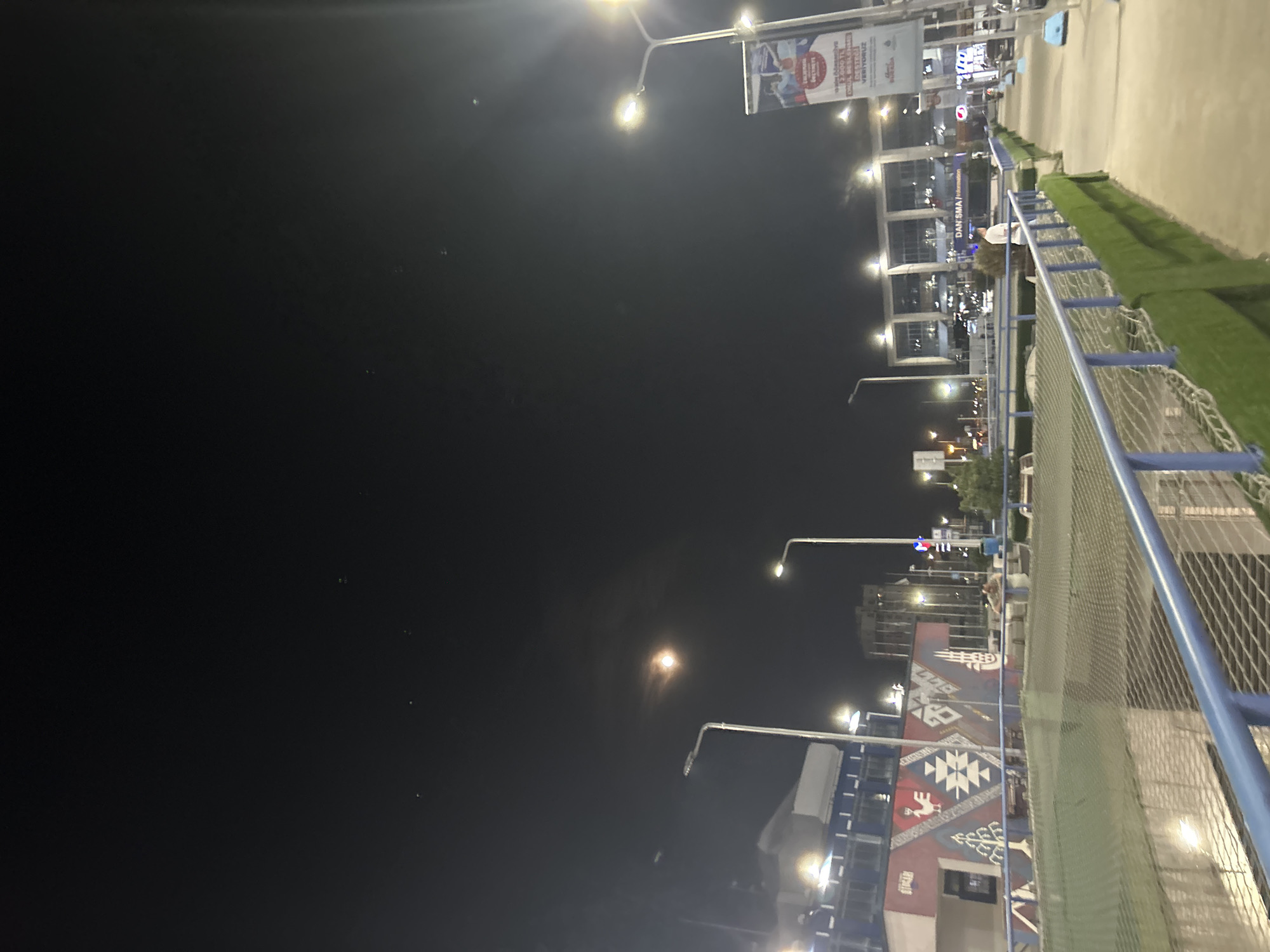 The image captures an urban night scene, likely in Bayrampaşa, Turkey, characterized by artificial illumination against a dark sky. A prominent bright light source, possibly the moon or a strong distant light, is visible in the upper-left sky, surrounded by numerous smaller, scattered bright points that appear to be distant lights or stars.
In the foreground, a blue metal railing or fence with a mesh pattern runs diagonally, separating a paved area from a strip of green artificial grass. Beyond this, several buildings with illuminated windows are visible. One building particularly stands out with a large, colorful mural on its side, featuring a geometric pattern reminiscent of traditional Turkish carpets (Kilim) in shades of red, white, and blue.
Numerous streetlights, equipped with bright lamps, line the street, casting strong glows and illuminating the surroundings. A large banner hangs from one of these streetlights. The visible text on the banner is in Turkish and reads: "Şimdi öğrencilere 2.500 TL burs desteği veriyoruz!" (Now we give 2,500 TL scholarship support to students!) and "DAHA İYİ GELECEK İÇİN BİRLİKTE YÜRÜYORUZ" (We walk together for a better future). The word "BÜYÜKŞEHİR" (Metropolitan Municipality) is also discernible, indicating the message likely comes from a municipal body. Another building has a sign that partially reads "DAN-SMA / information".
A single person is partially visible in the midground, standing near the fence, wearing a light-colored shirt or jacket. The time of day is clearly night, with the artificial lights providing the primary illumination. The scene depicts a quiet urban environment with commercial or public buildings.