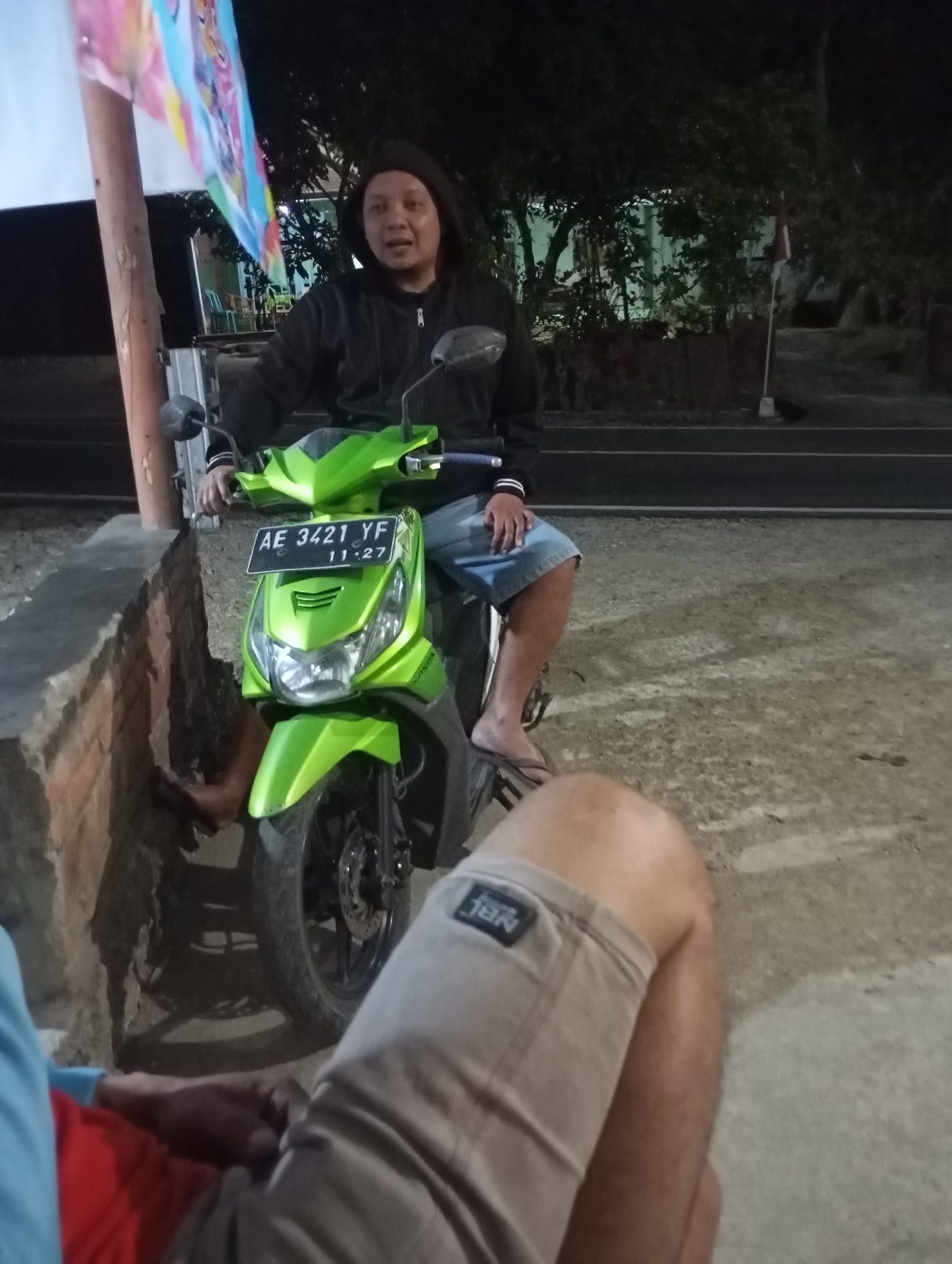 A man wearing a black hoodie and denim shorts is seated on a bright green scooter. He is looking towards the camera with an open mouth. The scooter has a license plate that reads "AE 3421 YF 11.27". The scene appears to be outdoors at night, possibly on a street in Wonogondo, Indonesia, given the presence of a flag pole with a flag and trees in the background. There is a low brick wall to the left of the scooter. Another person's legs are visible in the foreground, wearing tan shorts with a black tag.