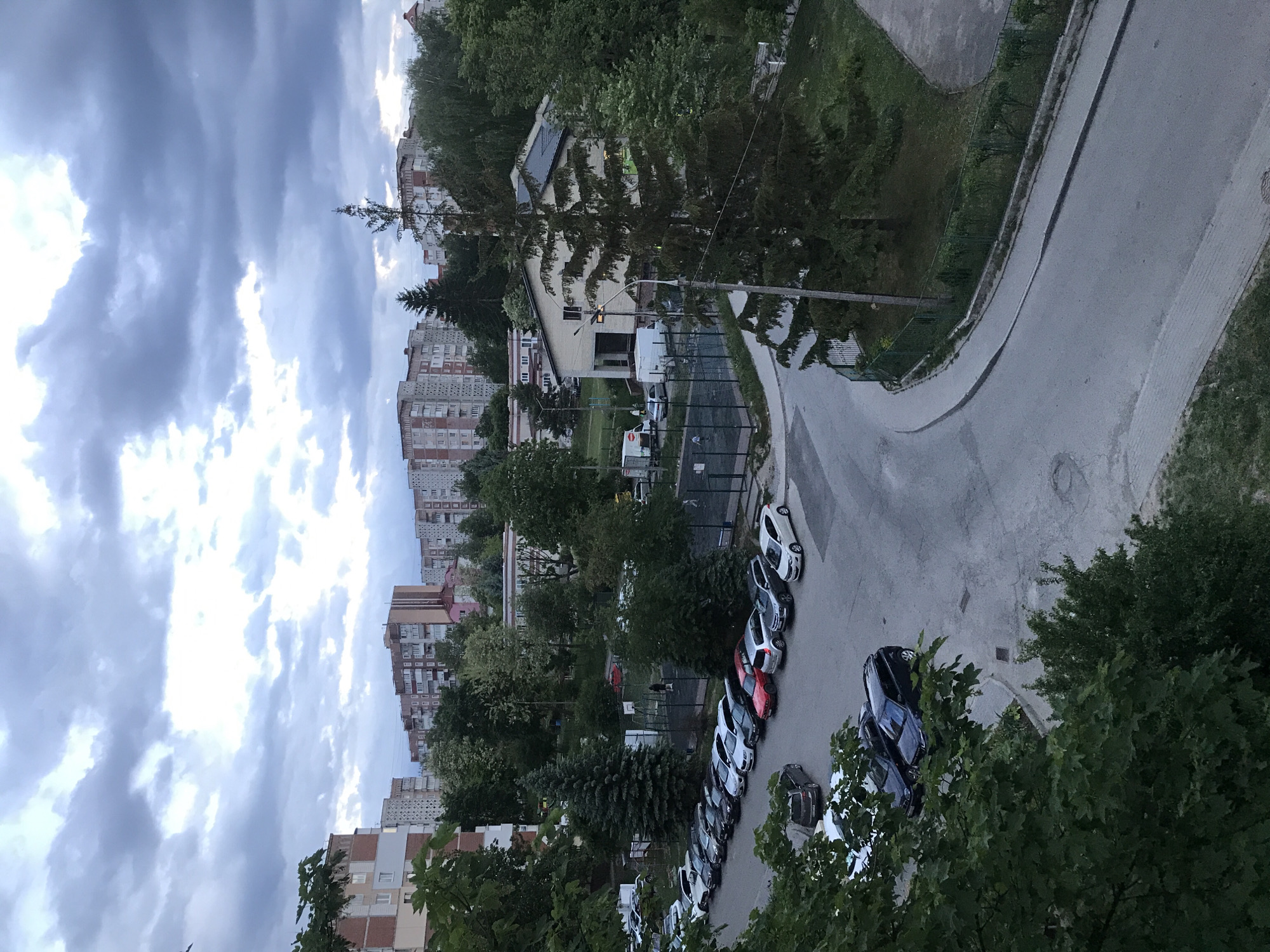 The image presents a view from an elevated position, overlooking a street with parked cars, buildings, trees, and cloudy skies. The scene is in Ternopil, Ukraine, evident from the architectural style of the buildings, which feature multi-story residential apartments, a common construction style found in Eastern European cities. A row of various colored cars is parked along the street. The street curves around a green area. Trees are interspersed among the buildings and line the street. The sky is filled with dense, grey clouds. There are no people visible in the immediate foreground. A fenced playground sits further back.