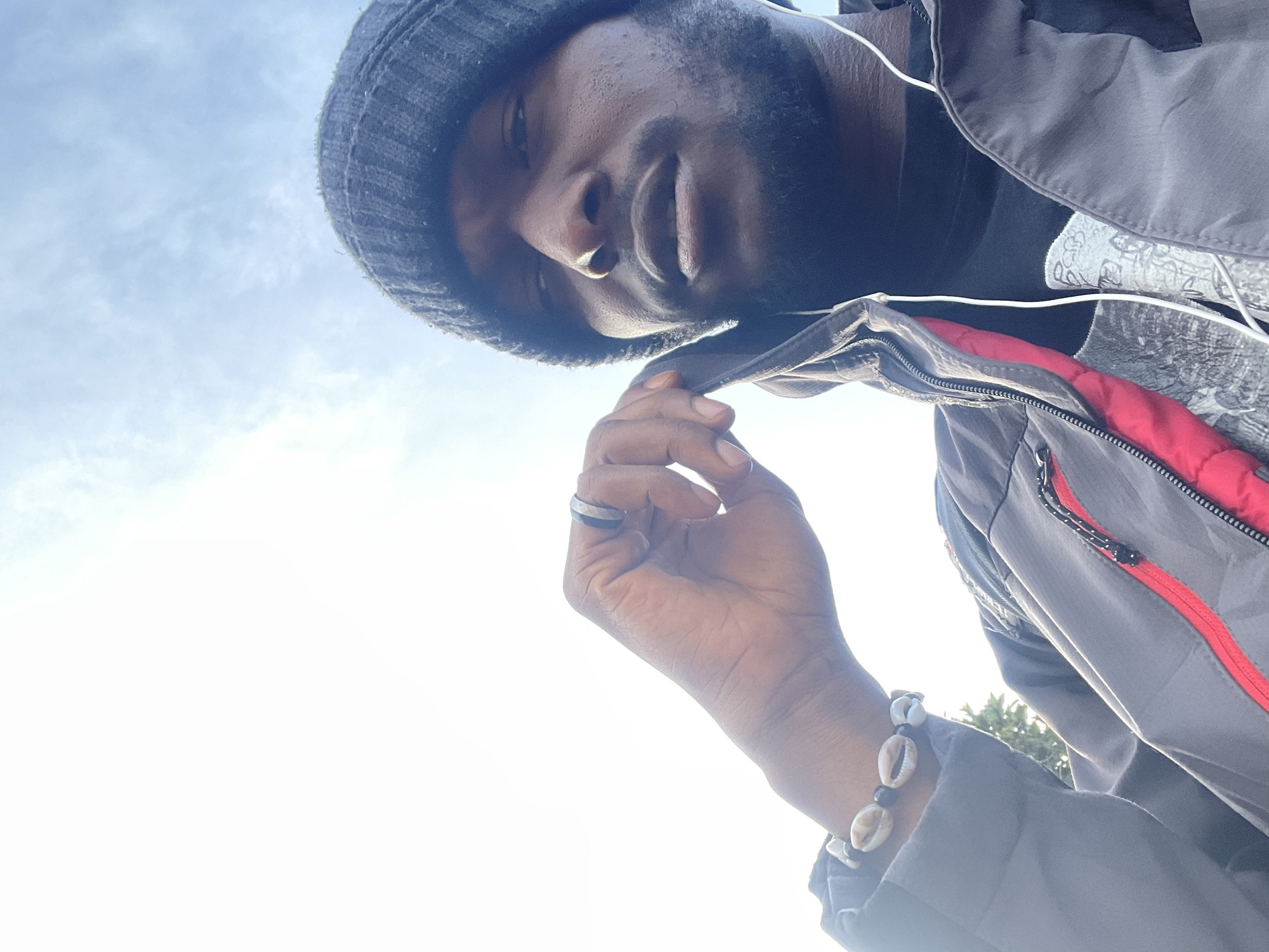 The image is a selfie taken from a low angle, looking up towards a person wearing a dark beanie, a grey and red hooded jacket, and white earbuds. The person has dark skin and a beard, and is wearing a ring on their left hand, which is resting near their face. A bracelet made of shells and beads is visible on their left wrist. The background is a bright sky with scattered clouds, indicating an outdoor setting. The image does not provide any specific visual cues to determine the location in Jos, Nigeria.