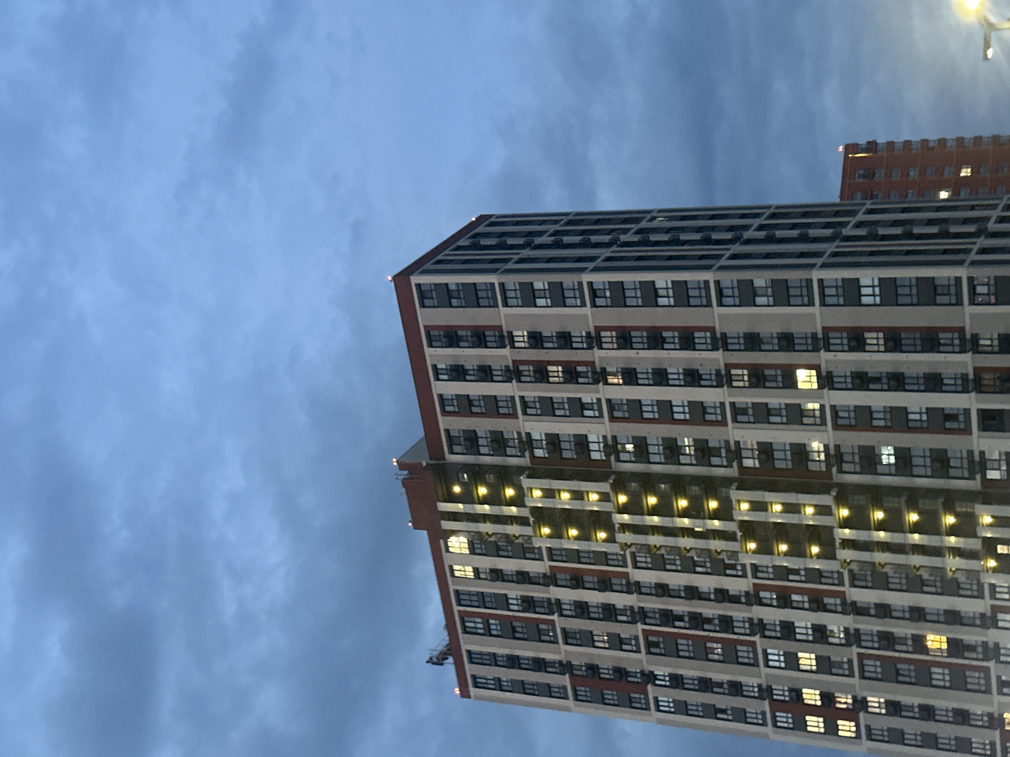 The image shows a tall, modern apartment building with a red-and-gray facade, set against a cloudy blue sky. The building has numerous windows, some of which are illuminated, suggesting that people are inside. A central vertical shaft runs up the building, featuring a series of yellow lights. On the roof of the building, a person is visible near the edge, and there are also small red lights on the roof's corners. To the right and slightly behind, a smaller, red brick building is visible, also with illuminated windows. A street light is partially visible in the bottom right corner. The scene appears to be captured at dusk or during a cloudy day.