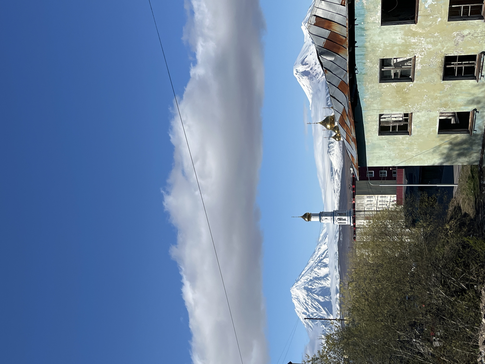 The image shows a view in Petropavlovsk-Kamchatsky, Russia. The left side of the image is dominated by a clear blue sky with a vertical strip of cloud. In the center, snow-capped mountains are visible, positioned behind a white church with a gold dome. To the right is a partially obstructed building with a rusty metal roof and several open windows. In the foreground on the right, there are leafy bushes. A power line runs down the left side of the image.