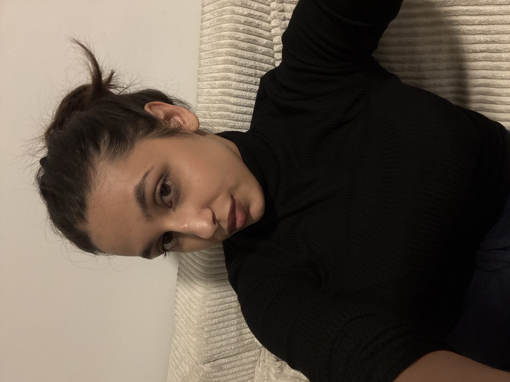A woman lies on a light-colored, ridged cushion or sofa. She has dark hair pulled up in a bun with a few stray strands. She wears a black, long-sleeved turtleneck sweater. Her head is turned towards the viewer, and she is looking at the camera. The background is a plain, light-colored wall. The location is uncertain. There is no clear evidence to confirm that this image was taken in the city of Mönchengladbach, Germany.