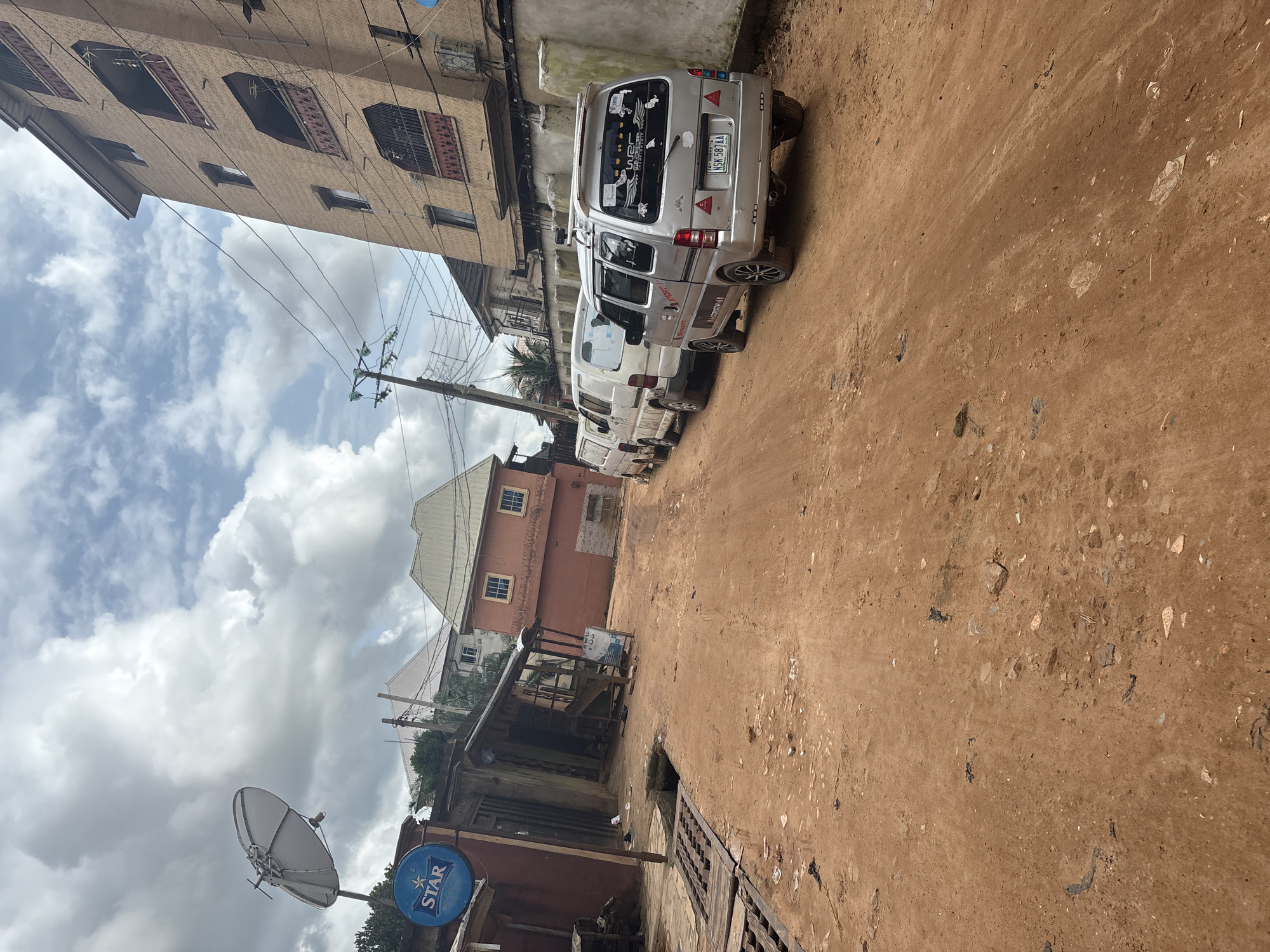 The image depicts a narrow, unpaved dirt road in Nkpor, Nigeria, lined with buildings. The road surface is a reddish-brown earth, with scattered debris, including plastic, rocks, and metal grates along the edges. A row of utility poles with tangled electrical wires runs along the right side of the road.
Several vehicles are parked on the right side of the road. The closest vehicle is a silver minivan with a Nigerian license plate (NSK 587AA) and decorative stickers on its rear window. Behind it are two white vans, also parked closely together.
On the left side of the road, a red building with a prominent satellite dish and a blue sign with the word "STAR" is visible. Adjacent to this building is a smaller, dilapidated structure with a tin roof, which appears to be a shop or stall. Further down the road, taller buildings with multiple stories and balconies are present, including a large tan building on the far right. The sky is partly cloudy, with a mix of white clouds and blue sky.