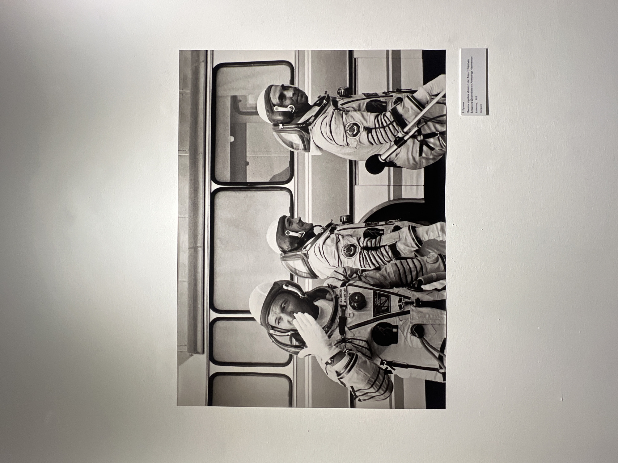 The image shows a black and white photograph hanging on a white wall. The photograph depicts three astronauts wearing space suits inside what appears to be a transport vehicle with rectangular windows. The text beside the photograph indicates the crew of the Soyuz T-6 spacecraft: Jean-Loup Chrétien, Vladimir Dzhanibekov, and Alexander Ivanchenkov at Baikonur in 1982. The location is not in Saint Petersburg, Russia, but at Baikonur Cosmodrome.
