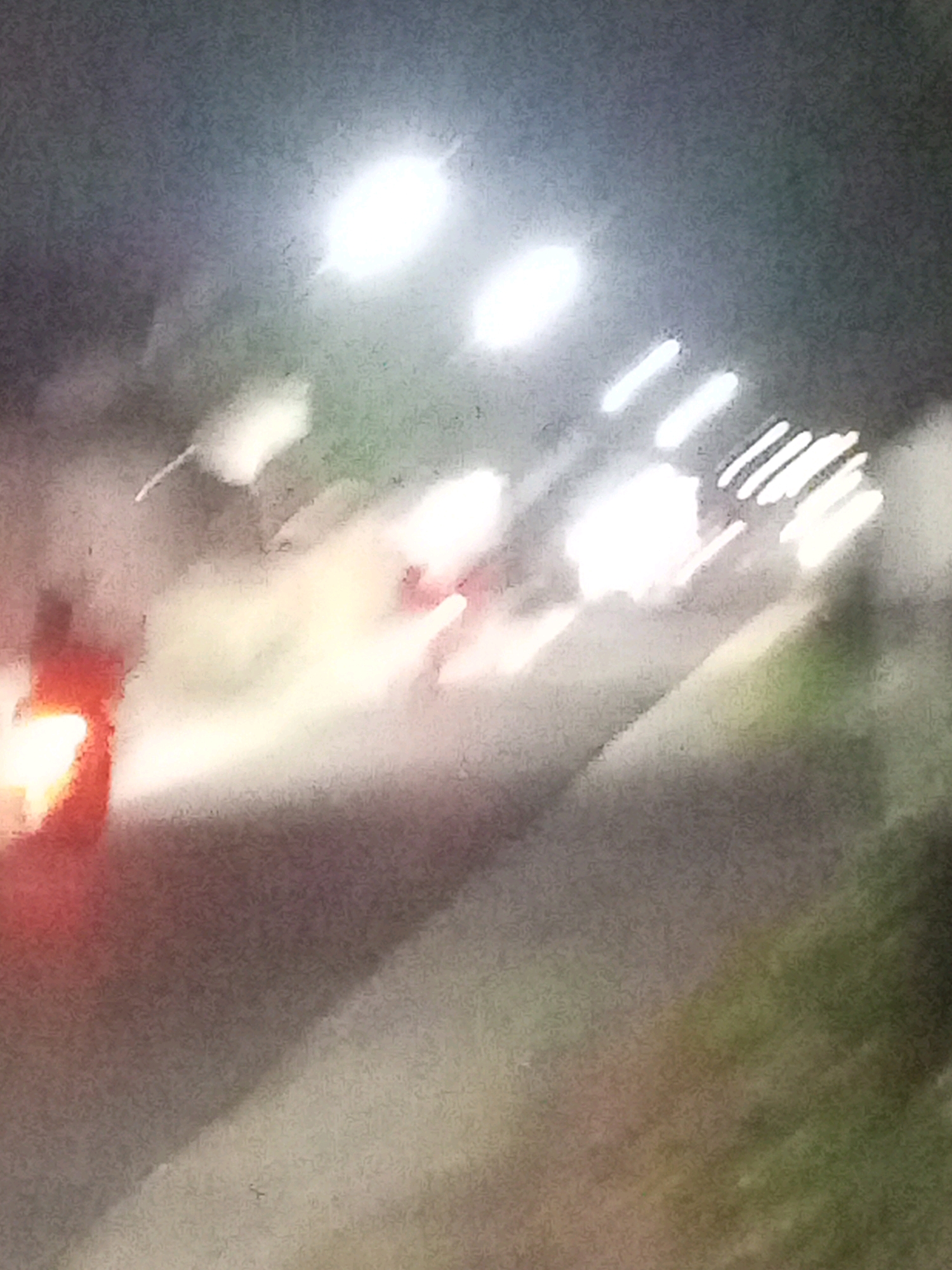 The image, captured at night in Ngasem, Indonesia, depicts a blurred scene of streetlights illuminating a road. A prominent red and white light source, possibly the taillights of a vehicle, is visible on the left. The overall impression is one of motion blur, with streaks of light suggesting activity on the road. The environment appears to be an urban or semi-urban setting, with the characteristic glow of street lighting. The lack of sharp detail makes it difficult to identify specific objects or individuals definitively.
