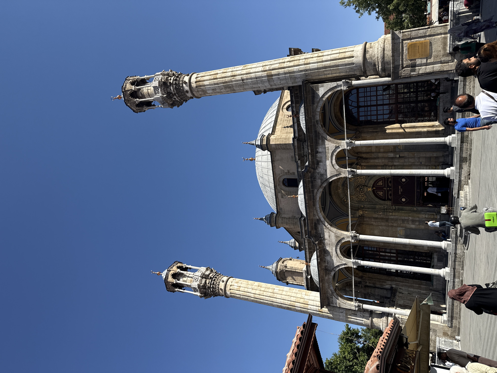 This is a low-angle, exterior, eye-level, full shot of the Aziziye Mosque in Karatay, Turkey, captured on a clear, bright day. The mosque features a central dome and is flanked by two tall, ornate minarets. The building's facade is characterized by a series of arched walkways supported by columns. People are visible in the foreground and near the entrance of the mosque, with some walking, others standing, and one person riding a scooter. The environment appears to be an open plaza or courtyard in front of the mosque, with glimpses of trees and parts of other structures to the sides, including a tiled roof on the left. The sky is a solid, deep blue.