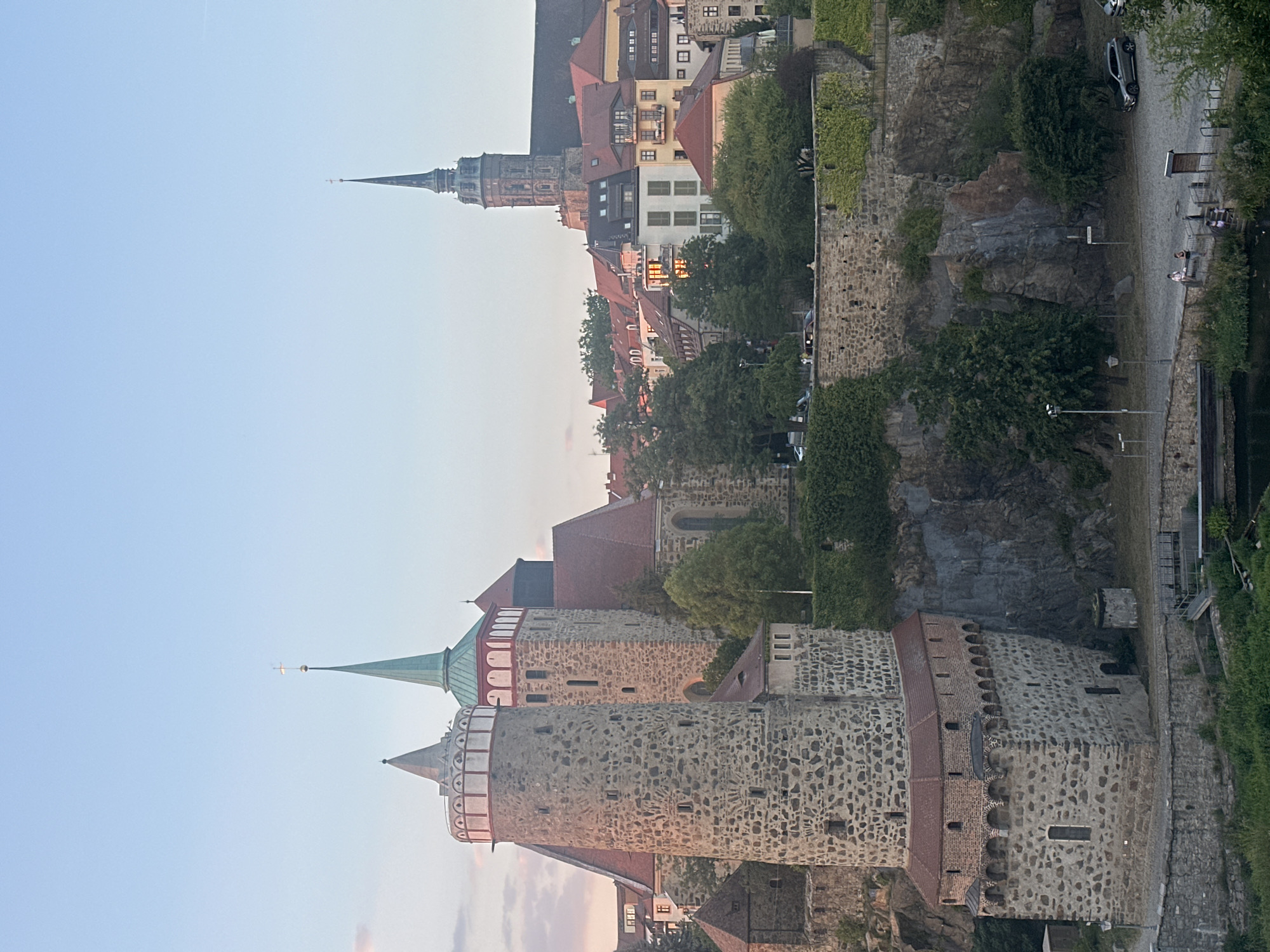 The image shows the Reichen Tower and the St. Peter's Cathedral in Bautzen, Germany, during the golden hour. The Reichen Tower, a prominent medieval structure made of stone, is on the left, featuring circular turrets and arched windows. Adjacent to it is St. Peter's Cathedral, with its distinctive green spire and red-tiled roof. In the background, a cityscape of traditional German architecture with red roofs and a tall church spire can be seen. The scene is set against a soft, pastel-colored sky. The foreground depicts a stone wall covered in ivy, a paved area with parked cars and a few pedestrians, and a body of water.