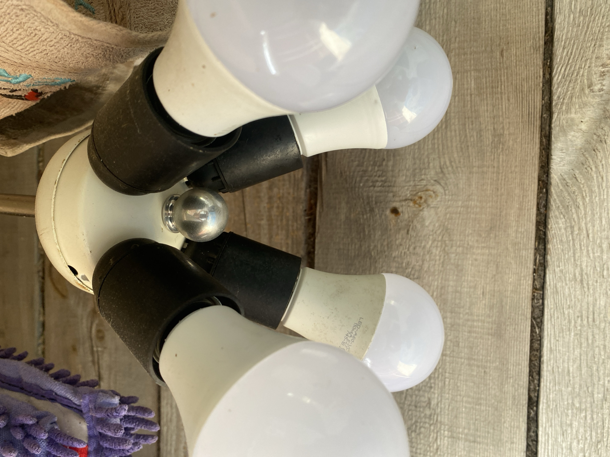 The image shows a close-up, low-angle view of a ceiling light fixture with four LED bulbs. The fixture is white with black accents around the bulb sockets. The bulbs are white and appear to be new. The light fixture is mounted on a wooden ceiling, which is made of planks. In the background, to the left, a purple and white duster is visible, and to the right, a tan towel with some embroidery is hanging. The overall impression is of an indoor or covered outdoor setting, possibly a porch or garage. The image does not contain any discernible features that would allow for the specific identification of its location within Krupki, Belarus.