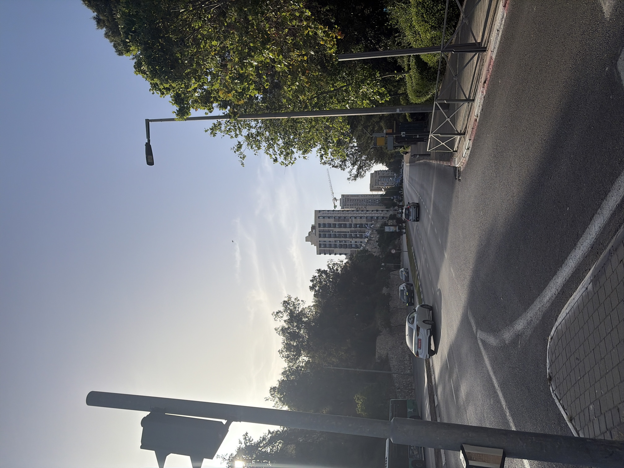 The image shows a street view in an urban area, likely in Unknown, Unknown. The road curves slightly to the right, with several cars parked along the side. Buildings and trees are visible in the background. A construction crane stands near the buildings. A street light is on the left side of the road. The sky is clear.