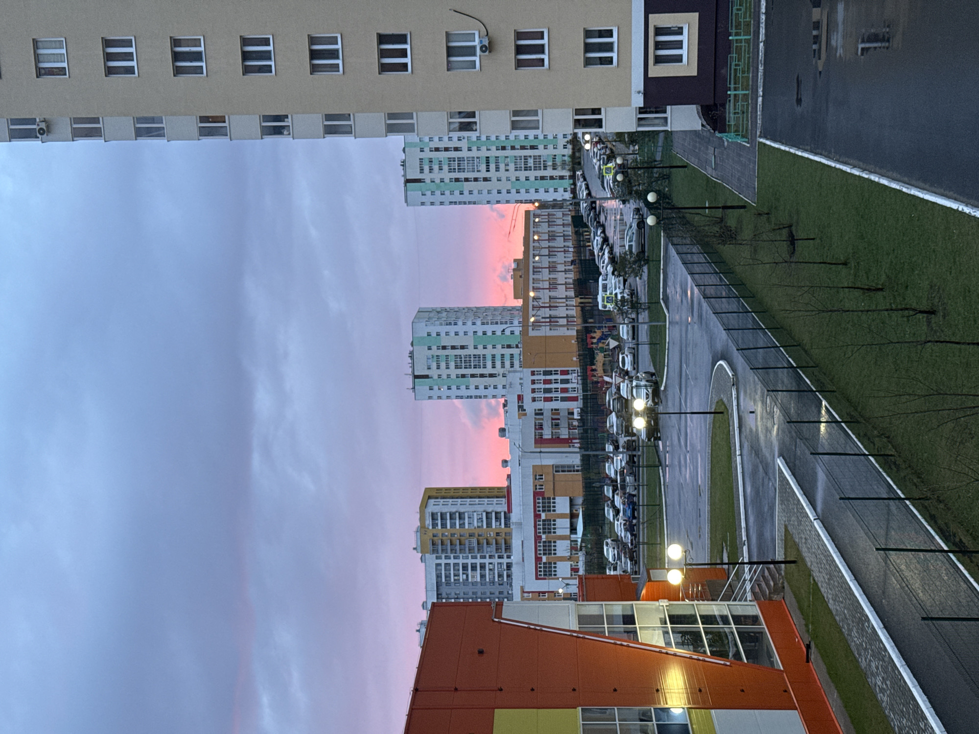 The image shows a view from an elevated position of a residential area, presumably in Tobolsk, Russia, given the architectural style common in Russian cities. Multiple high-rise apartment buildings are visible in the foreground and mid-ground. The buildings are of various colors, including shades of beige, green, orange, and white, and display a repetitive window pattern. The sky is overcast with pink hues suggesting either dawn or dusk. A parking lot filled with cars and illuminated by street lamps is situated between the buildings. A sidewalk lined with a chain-link fence and saplings runs alongside the parking area. The lighting conditions appear dim and slightly wet. The composition suggests a dense urban environment.