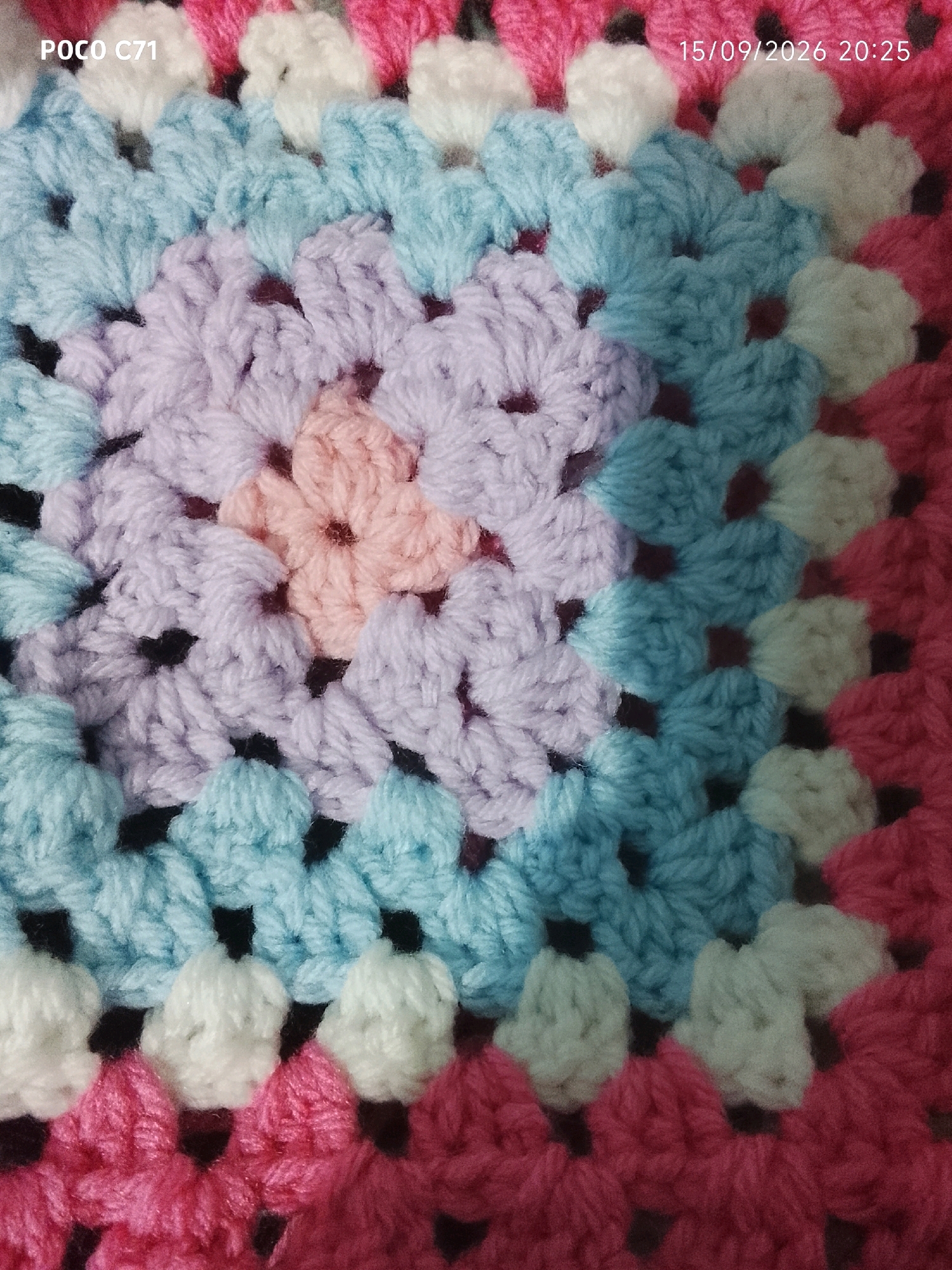 The image is a close-up, high-angle shot of a crocheted textile. The textile features a repeating hexagonal pattern, commonly known as a granny square, constructed with yarn in various pastel colors including pink, white, light blue, and lavender, with some darker stitches visible between the larger crocheted sections. The lighting suggests an indoor setting, with a timestamp in the upper right corner reading "15/09/2026 20:25" and "POCO C71" in the upper left. There are no people or identifiable landmarks present. The image does not contain any information that allows for the determination of its location in Bhateliya, India.