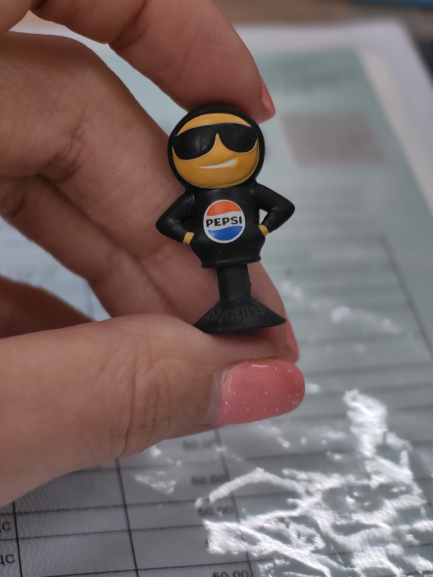 A miniature figurine of a yellow smiley face wearing sunglasses and a black hoodie with a Pepsi logo is held by a person's hand. The figurine has its hands on its hips. The background appears to be a blurry document with grid lines and some text. The location is not specified in the image.