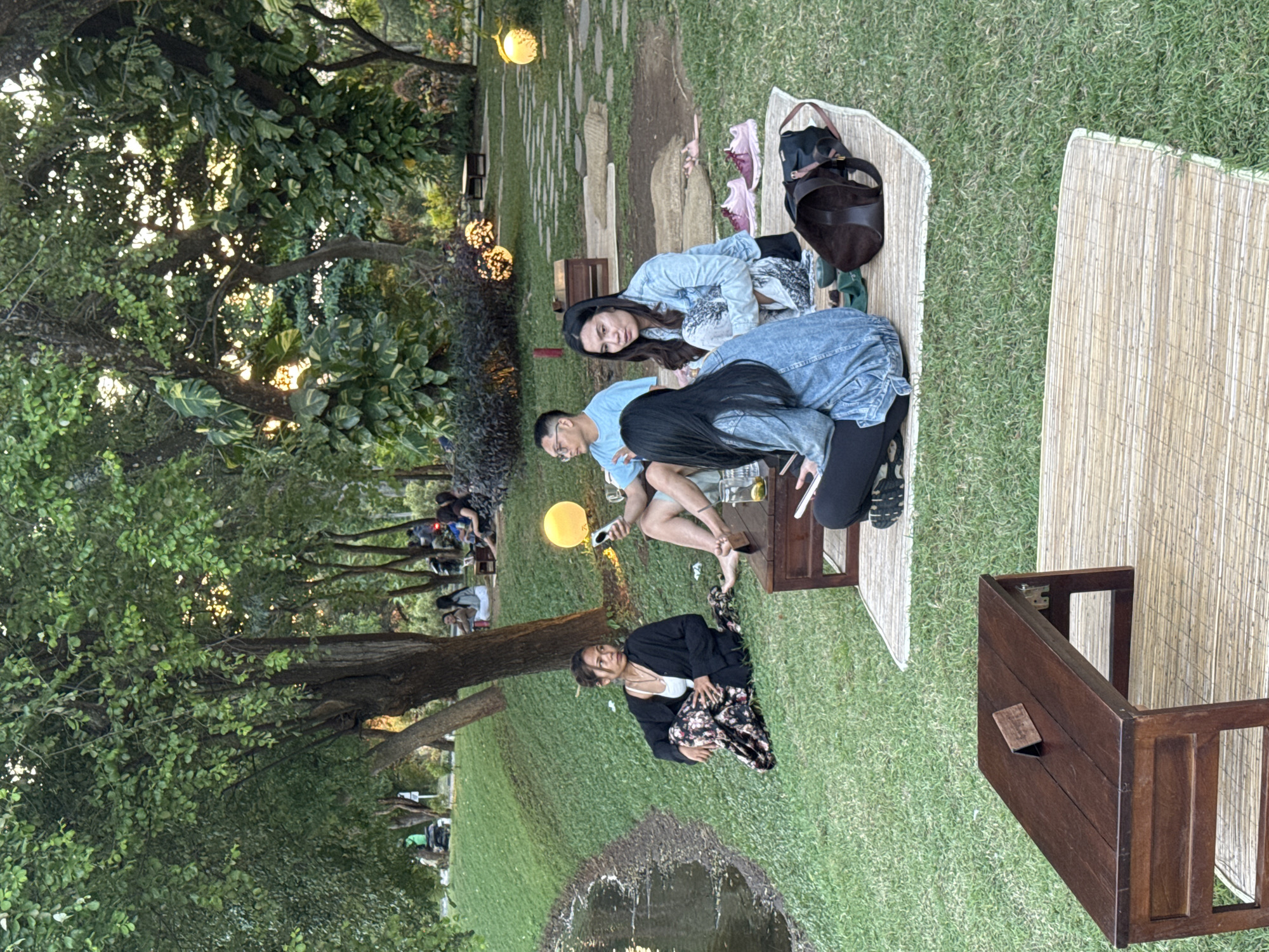 A group of three young adults are sitting on a picnic blanket in a park-like area, near Pecatu, Indonesia. The group includes two young women and one young man. The picnic setup includes a small wooden table, bags, and other picnic essentials. One of the women is kneeling while the other two are sitting near the table. In the background, there is a group of people sitting at a picnic area, and in the foreground, there is a small water feature. There are trees on the left side of the scene and string lights hanging above the area. A picnic table sits off to the side.