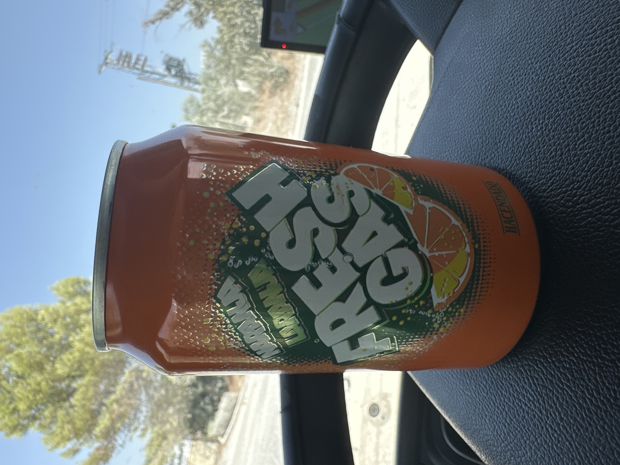 An orange soda can, labeled "NARANJA LORANHA FRESH GAS," sits on a black, textured surface, likely a car's dashboard. The can features green graphics with orange slices and white lettering. In the background, a bright green tree is visible against a clear blue sky, with some power lines and a blurry vehicle interior suggesting an outdoor, possibly roadside setting. The label "HACENDADO" is visible on the lower portion of the can. The scene is well-lit, indicating daytime.