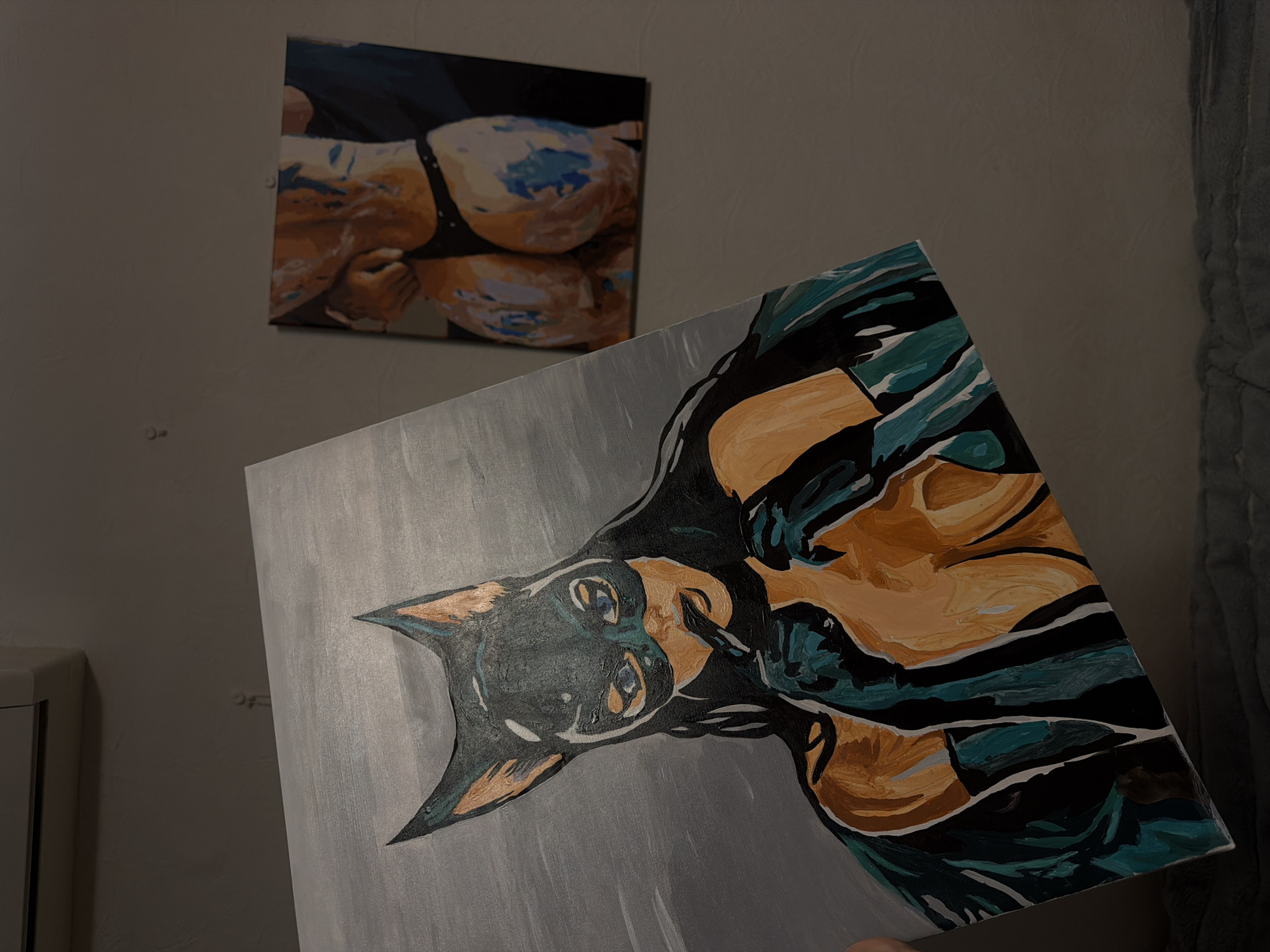 The image presents an indoor scene, dimly lit, which according to the provided context is located in Moscow, Russia. The main subject in the foreground is a canvas painting, held at an angle, depicting a stylized Doberman Pinscher. The dog's head is predominantly black and dark brown with prominent blue eyes, set against a light grey, brush-stroked background. Its body is abstractly rendered with bold, sweeping strokes of orange, teal, and black, suggesting movement or form.
In the background, mounted on a neutral-colored wall, is a second painting. This artwork portrays a close-up section of a human torso, specifically the upper chest and breasts, wearing a black garment resembling a bra or bikini top. The skin is depicted in warm brown and orange tones, accented with patches of light blue paint, possibly indicating reflections or artistic abstraction.
To the far left of the frame, a portion of a light-colored cabinet or piece of furniture is visible. On the far right, a grey, textured fabric, likely a blanket or curtain, adds to the domestic setting. The wall behind the paintings is unadorned and a uniform light tone. The activity captured is the display or viewing of these two pieces of art. There is no visible text in the image.