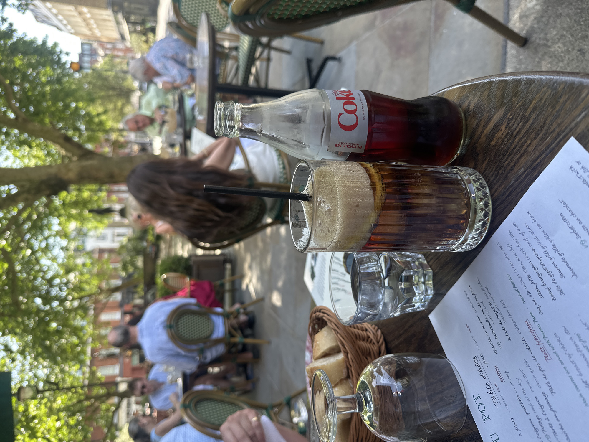 Here is a description of the image:
**Scene:** The image captures a scene at an outdoor restaurant, possibly in Europe, with a focus on a table setting. It's daytime, and the setting appears to be a bustling area, suggested by the presence of numerous people in the background.
**Objects:**
* A partially filled glass of what looks like a dark soft drink with foam. A black straw is visible in the glass.
* A glass bottle of Coca-Cola, with the label showing "Coke, No Sugar No Calories."
* Two other empty glasses.
* A basket of bread.
* A menu, displaying a French menu with "Table d'Hôte" (set menu) and dishes listed in French.
**People:**
* Multiple people are sitting at other tables. There are people of various ages.
* The edge of a person's hand is visible at the bottom left of the frame.
**Environment:**
* The restaurant appears to be located outdoors, with trees and buildings visible in the background.
* The tables and chairs are typical of a cafe or bistro setting.
**Overall:** The photograph captures a candid moment at an outdoor restaurant, highlighting the drinks and the presence of diners enjoying a meal.