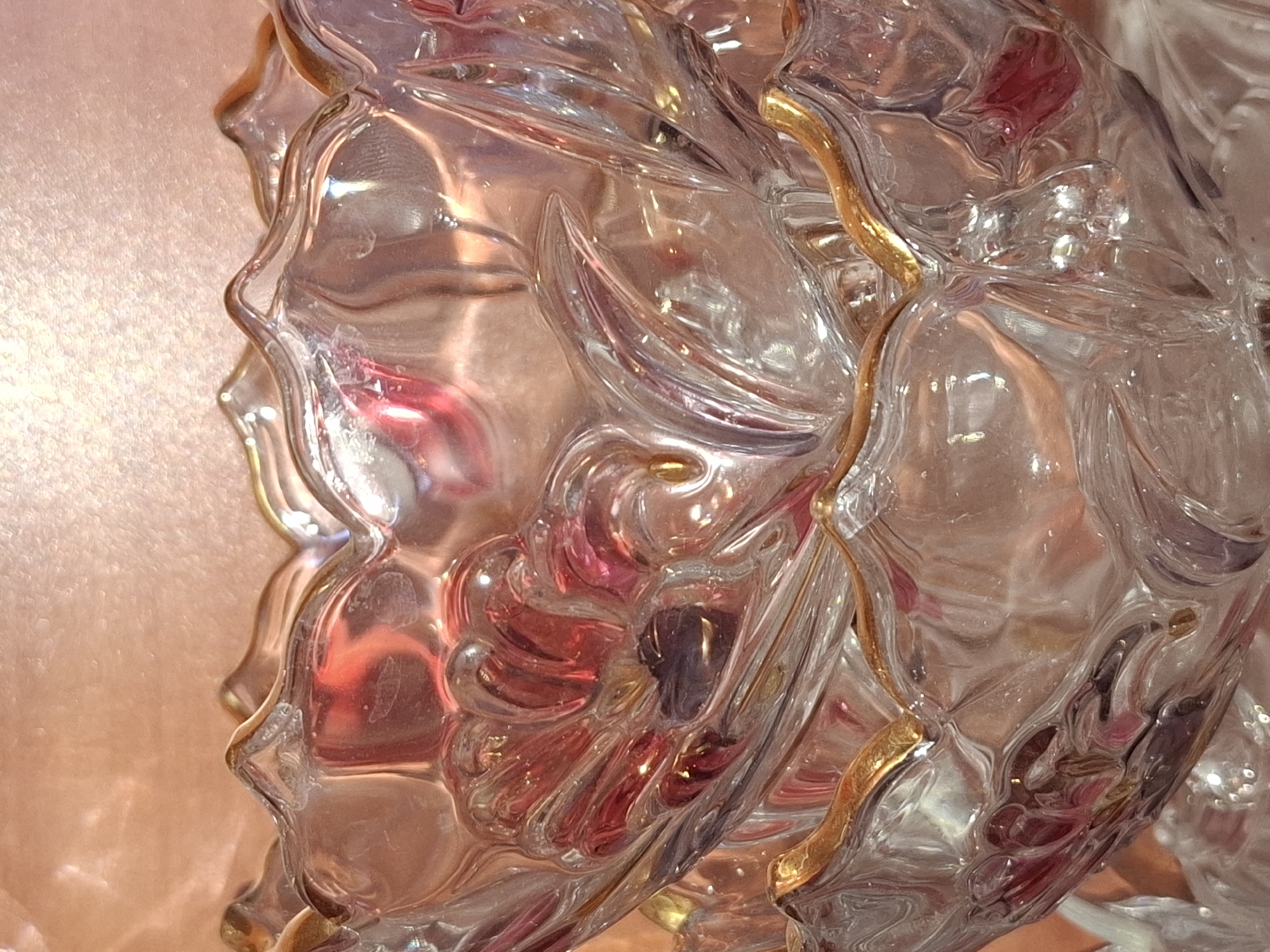 The image displays a close-up view of two stacked glass bowls, intricately designed with floral or petal-like embellishments. The glass appears clear with a subtle pinkish or rose-gold hue, reflecting light and creating a shimmering effect. Gold trim outlines the wavy edges of the bowls. The background is a textured, soft pink surface that appears to be fabric or a similar material, out of focus. The scene is static and focuses solely on the decorative objects. There are no people, animals, or specific environmental features identifiable as being in Almaty, Kazakhstan.