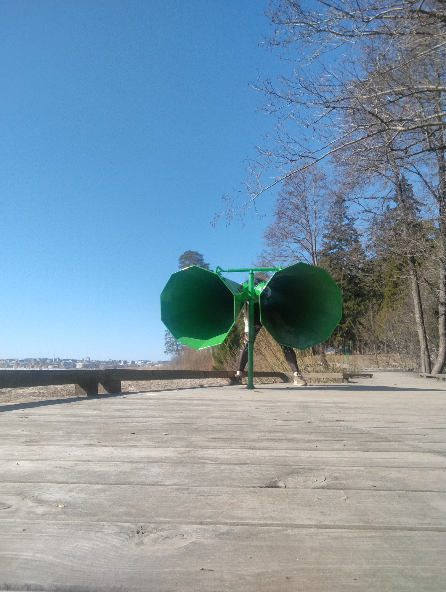 The image shows a scene in Votkinsk, Russia. A person is standing behind a large, green, double-horned sound sculpture positioned on a wooden boardwalk. The sky is clear and blue. A low cityscape is visible in the distance. To the right, a wooded area with bare trees is present. The boardwalk appears to be elevated, with a wooden railing visible in the mid-ground. The person is wearing dark leggings and light-colored sneakers, with their legs visible between the two green horns.