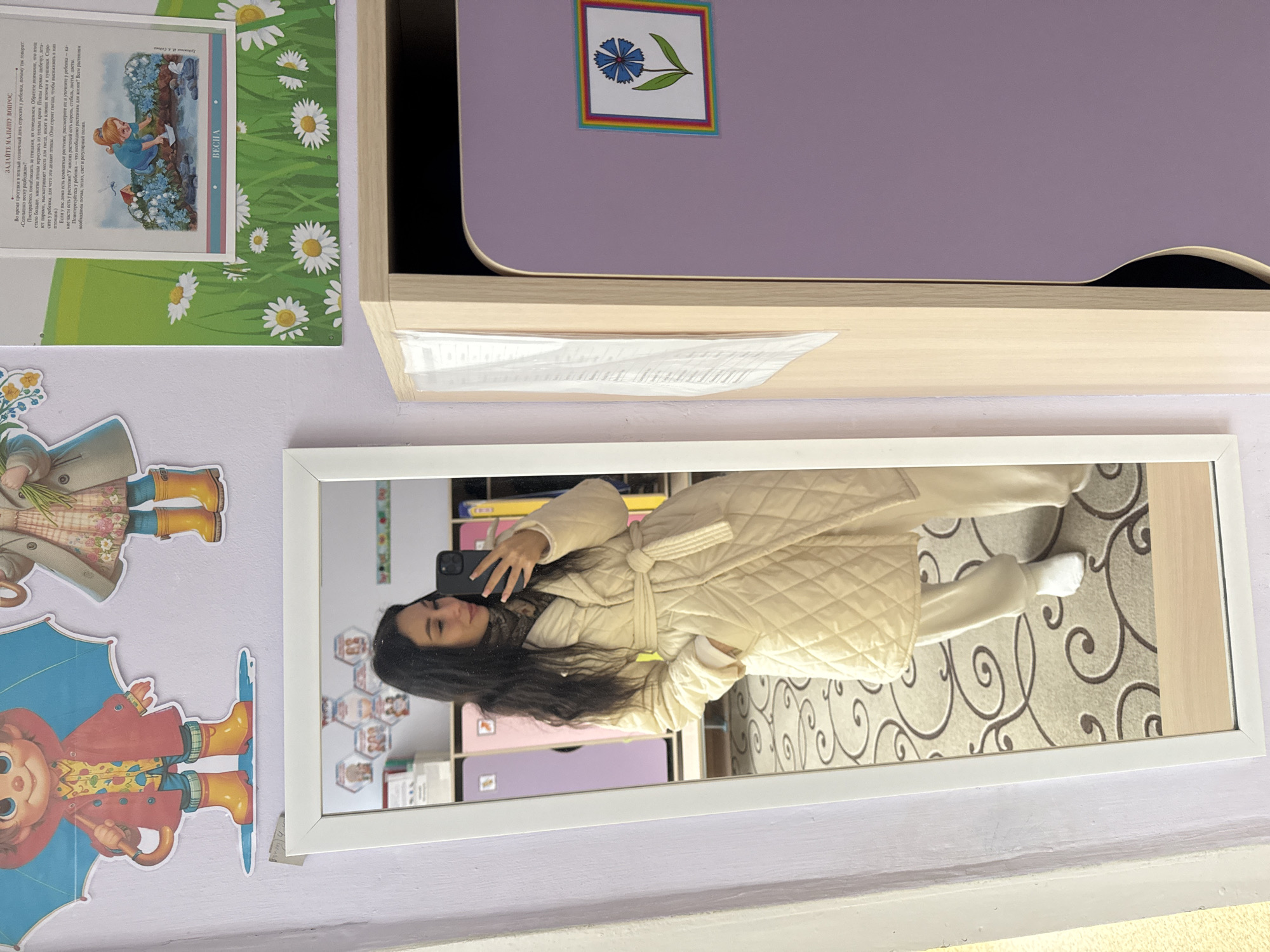 Here's a detailed description of the image:
The photograph appears to be a selfie taken in a room. The subject, a person with long dark hair, is reflected in a rectangular mirror with a white frame. They are wearing a light-colored, quilted robe-like garment with a tie at the waist and white socks. They hold a smartphone in their hand, presumably to capture the reflection.
The mirror reveals a room with shelves filled with items. The walls are a light color, possibly lilac or lavender. On the wall to the left of the mirror, there are decorations or stickers, including what appears to be a child-like illustration of a figure with an umbrella and boots.
Above the mirror, there is a shelf or desk area that is also light in color, topped with a purple surface with a flower decal. A printed sheet with text and illustrations is taped to the upper-left corner of the image.
The location seems to be in a room, likely a child's bedroom or a classroom based on the decorations. The OCR text "ЗАДАЙТЕ МАЛЫШУ ВОПРОС" is in Russian and the area might be in the city of Chaikovsky, Russia.