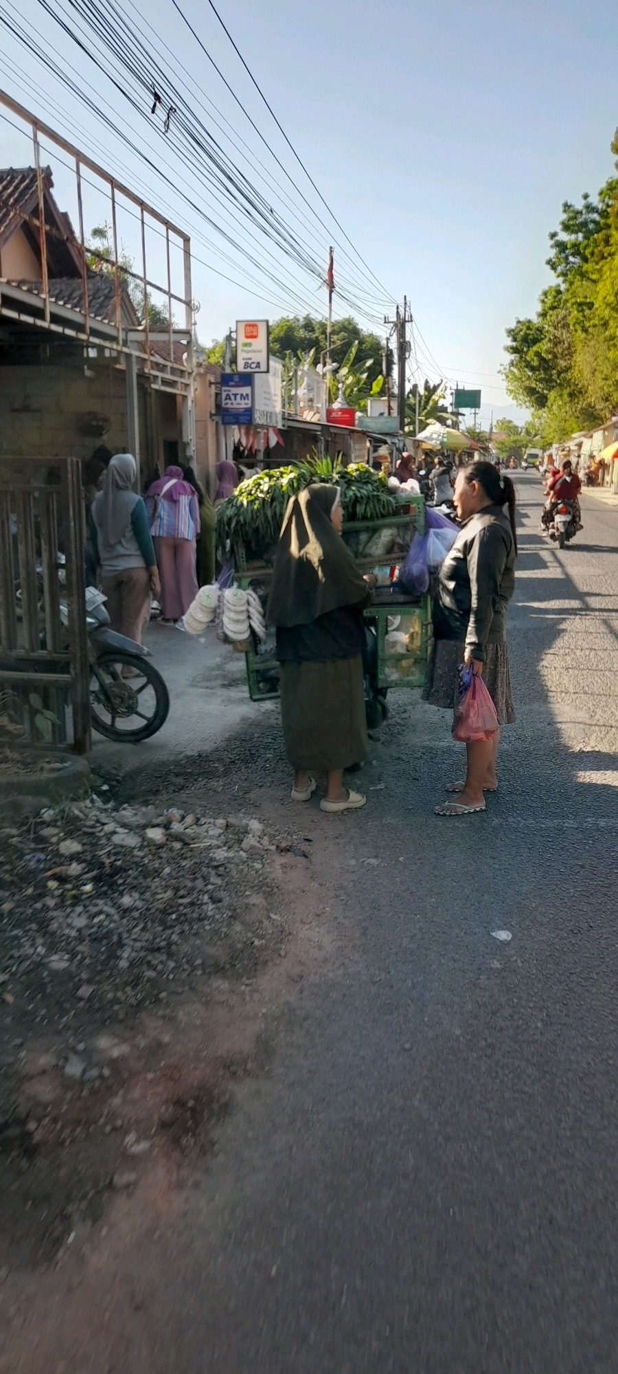 In Kendalsari, Indonesia, a street-side vegetable cart is the focal point of a scene. Two women are engaged in what appears to be a transaction or conversation at the cart. The woman on the left, wearing a dark headscarf and a long skirt, is facing the cart. The woman on the right, in a dark jacket and patterned skirt, is facing the woman on the left and holding a pink plastic bag. The cart is laden with fresh produce, predominantly green leafy vegetables. Behind the cart, other individuals and a motorbike are visible, suggesting a bustling market environment. To the left, a building with an open entrance and a parked motorbike further detail the setting. Overhead, a complex network of power lines stretches across the sky. The ground is paved with asphalt, with some debris and patches of dirt along the side of the road. The lighting suggests it is daytime, with sunlight casting shadows.