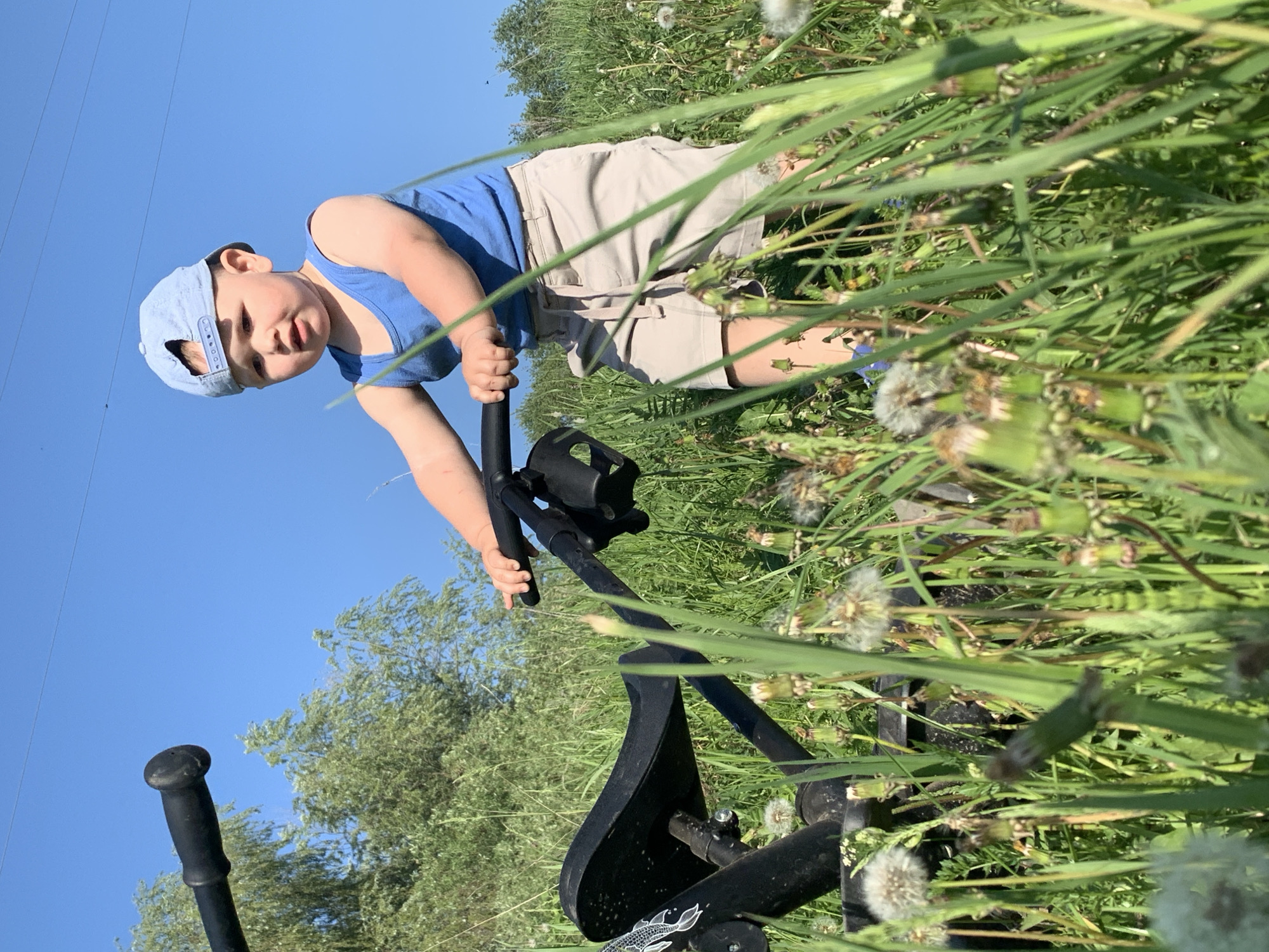 A child is standing in a field of tall grass and dandelions in what appears to be daylight. He is wearing a light blue baseball cap, a blue t-shirt, and khaki shorts. The child is leaning forward, gripping the handle of a black metal object that appears to be a piece of equipment of some kind, possibly a weed puller or similar tool. The sky is blue, and trees are visible in the background. It's not possible to confirm the location is Фоки, Russia based on visual information alone.