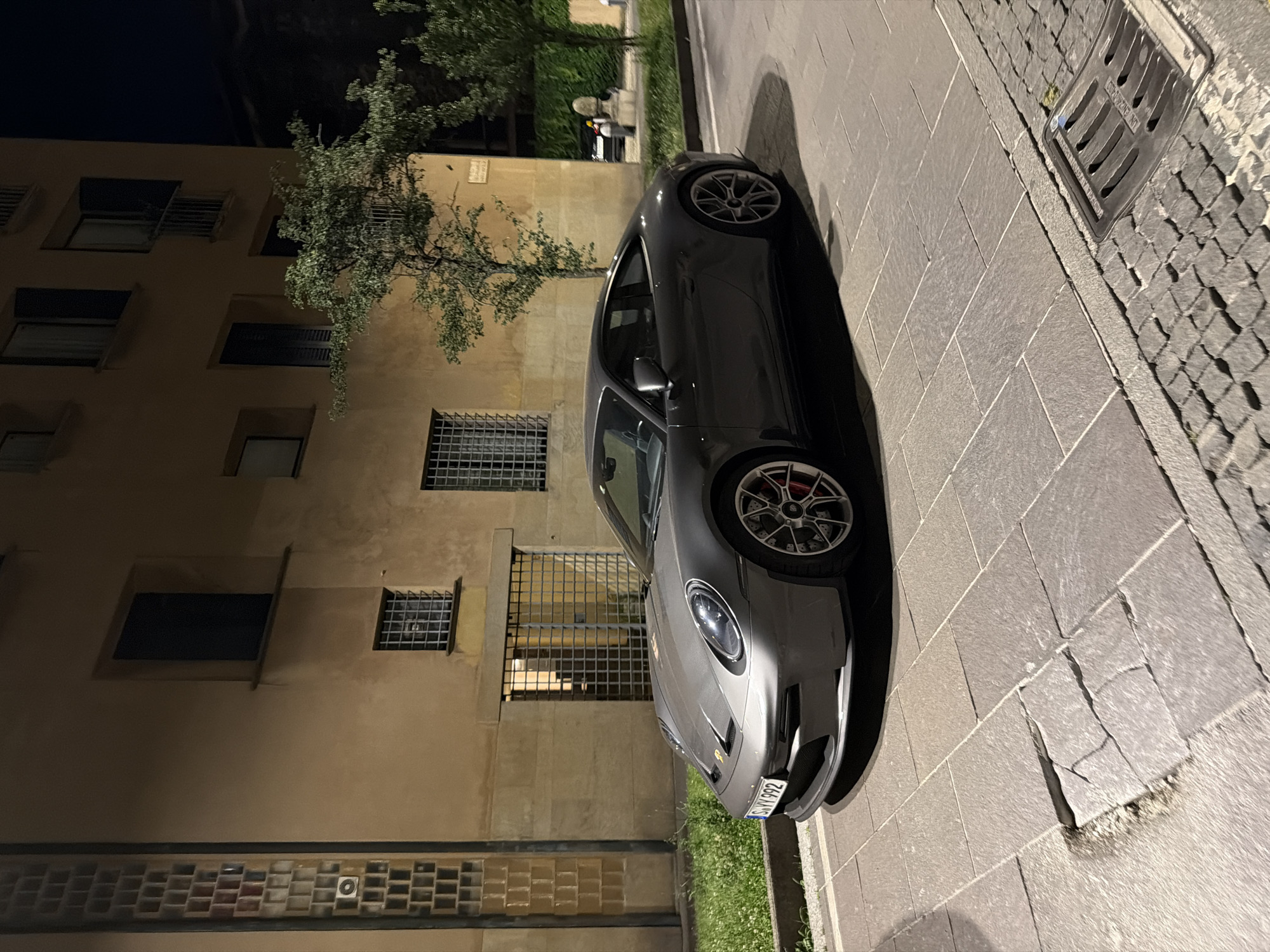 The image shows a gray Porsche 911 parked on a cobblestone street at night. The car is in good condition and appears to be a sports car. The background features a building with yellow stucco walls and several windows, some with grates. A tree with green foliage is positioned in front of the building. The street is paved with large, rectangular stones and there is a manhole cover on the right side of the image. The overall setting appears to be an urban environment, possibly in Milan, Italy, given the architectural style. There are no people clearly visible or interacting with the car or the environment. The lighting suggests it is nighttime, with artificial lights illuminating the scene.