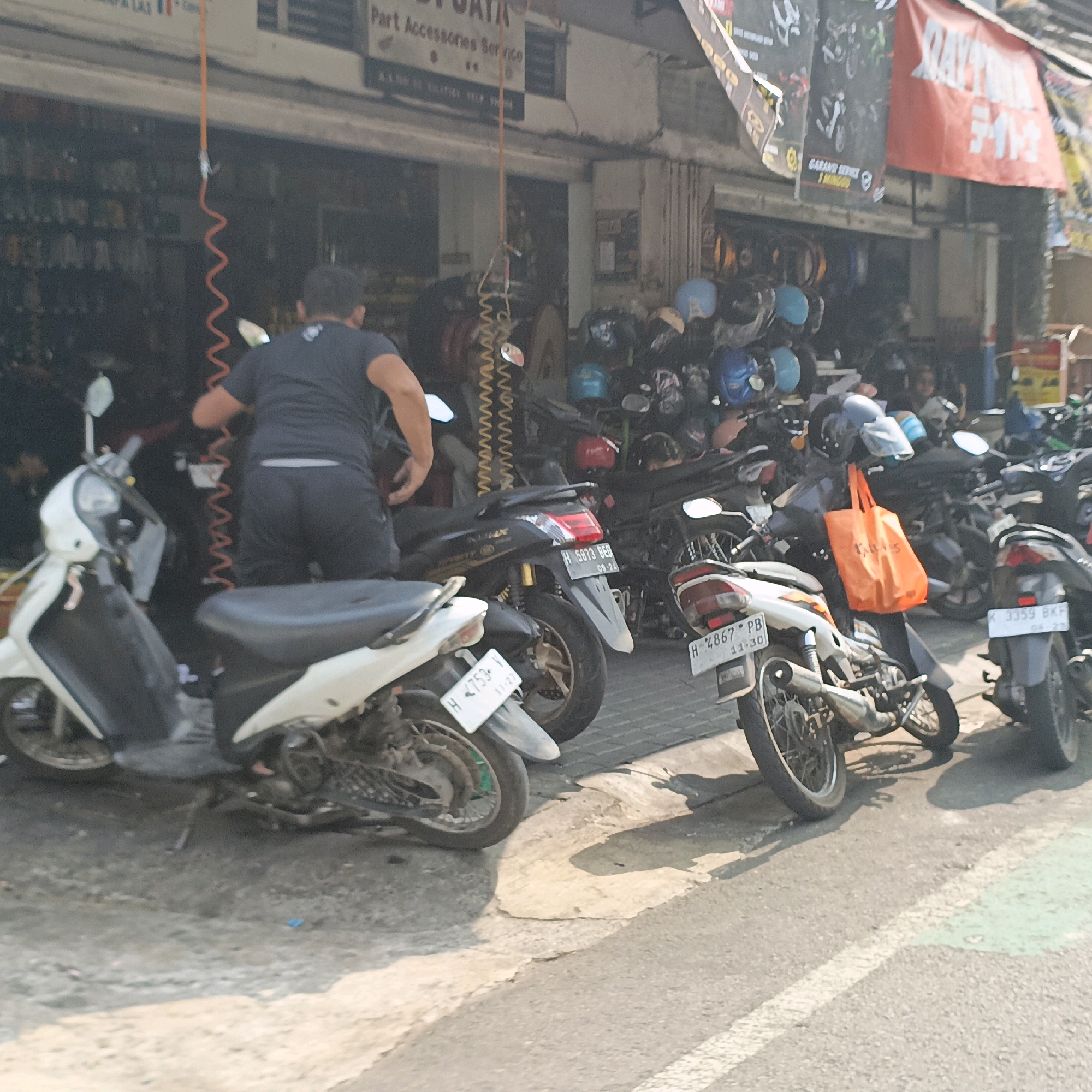 A street view captures a busy motorcycle repair shop in Salatiga, Indonesia, filled with parked motorcycles and a variety of helmets displayed. A person, wearing a dark t-shirt and shorts, is attending to a white scooter on the left. Another scooter, black with red taillights and a license plate reading "H 5873 BED", is parked to its right. A third scooter, also white with a license plate "H 4753 - 11-22", is in the foreground. Further to the right, a motorcycle with a bright orange bag hanging from its handlebars is visible, accompanied by another scooter with license plate "H 4867 PB 11-30". Additional motorcycles and helmets are observed in the background, suggesting a lively commercial area. The street is paved, with some concrete sections and tire marks visible. The shop itself appears to be a narrow building with an open facade, displaying various products.
