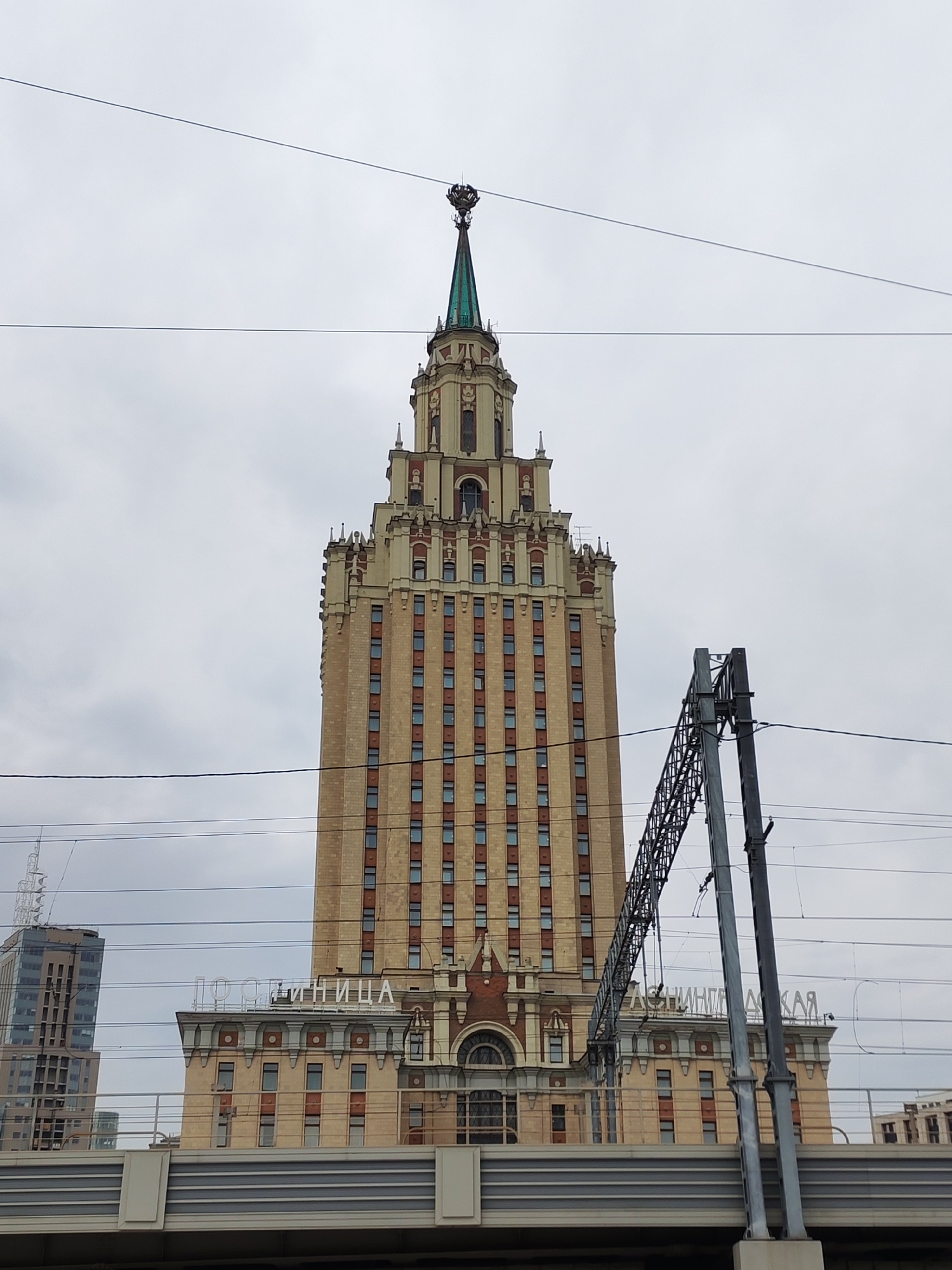 The image presents a tall, ornate building with architectural features reminiscent of Stalinist Empire style, situated in an urban setting under an overcast sky. The building dominates the center, constructed from light-colored brick or stone with red trim and numerous rectangular windows. It rises in a series of tiered sections, culminating in a distinctive green spire topped with an elaborate, crown-like ornament.
Several thin, dark power lines or cables crisscross the frame, partially obscuring the upper sections of the building and the sky. To the right, a dark gray metal lattice structure, likely part of an overhead railway or metro support, also intrudes into the view. In the foreground, a solid, gray bridge-like structure spans horizontally across the lower part of the image.
On the lower facade of the main building, large white Cyrillic letters are visible. On the left, the word "ГОСТИНИЦА" (GOSTINITSA), meaning "HOTEL," is clearly legible. To its right, partially obscured by the metal support, are fragments of what appears to be "ЛЕНИНГРАДСКАЯ" (LENINGRADSKAYA), identifying the structure as the Leningradskaya Hotel in Moscow, Russia.
To the far left in the background, a modern high-rise building with a glass facade is partially visible. The scene is static, devoid of people or discernible activity, and captures the building on a cloudy day, suggesting a daytime shot with diffused light.